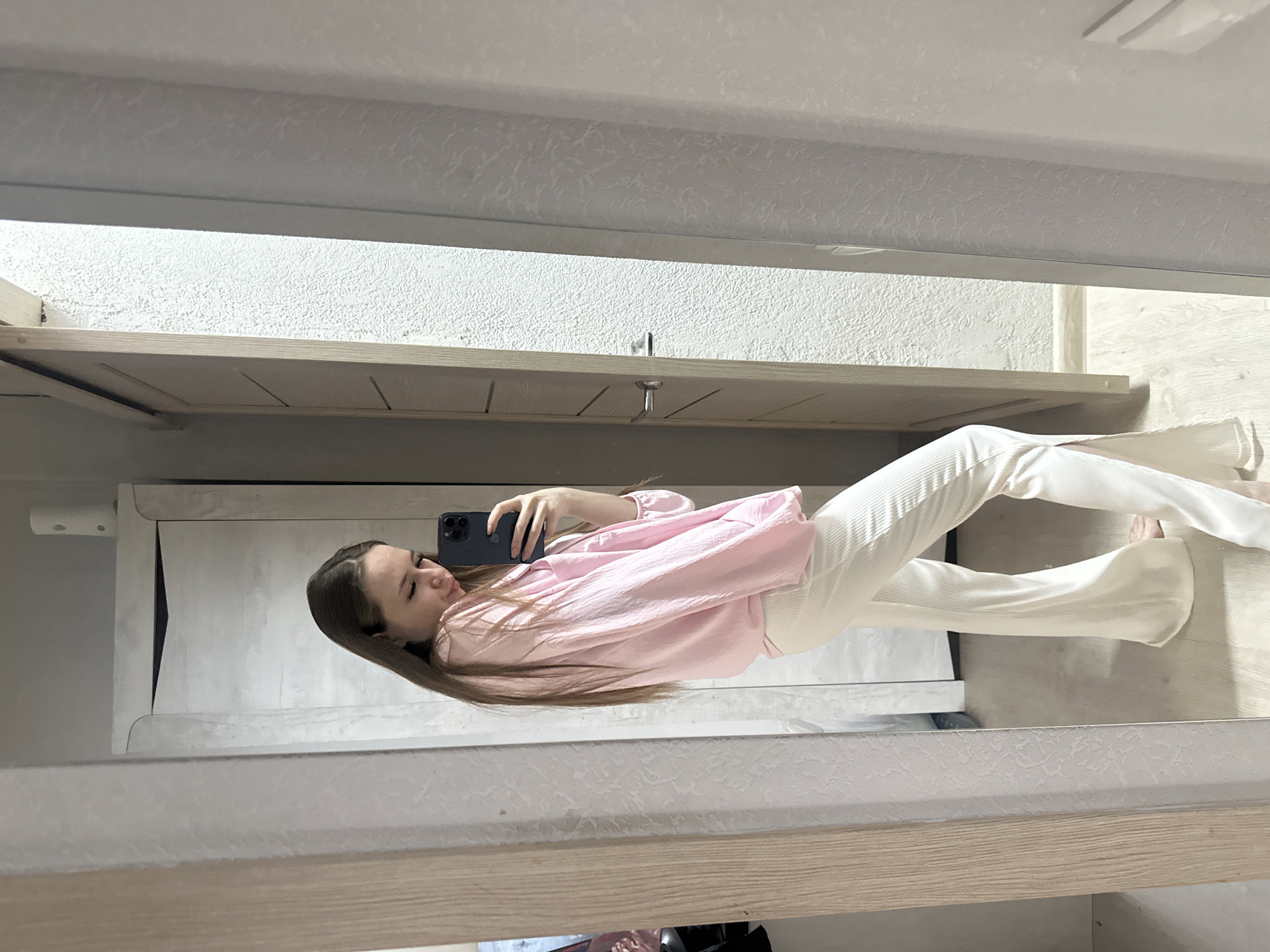 A young woman is captured in a mirror selfie, standing indoors. She is holding a dark-colored smartphone with multiple cameras in her right hand, positioned to take a photo of her reflection. Her long, dark brown hair cascades over her shoulders and down her back.
She is wearing a light pink, long-sleeved blouse with a loose fit and a textured or wrinkled fabric. Her bottom half is dressed in cream or off-white wide-leg pleated pants. Her expression is neutral as she looks towards the phone.
The setting, visible in the mirror's reflection, appears to be a room with light-colored, possibly textured, walls. There are light wooden elements, which seem to be part of a door, a cabinet, or a mirror frame. The floor reflected beneath her feet is also light-colored. The lighting suggests daytime. No text is visible in the image.