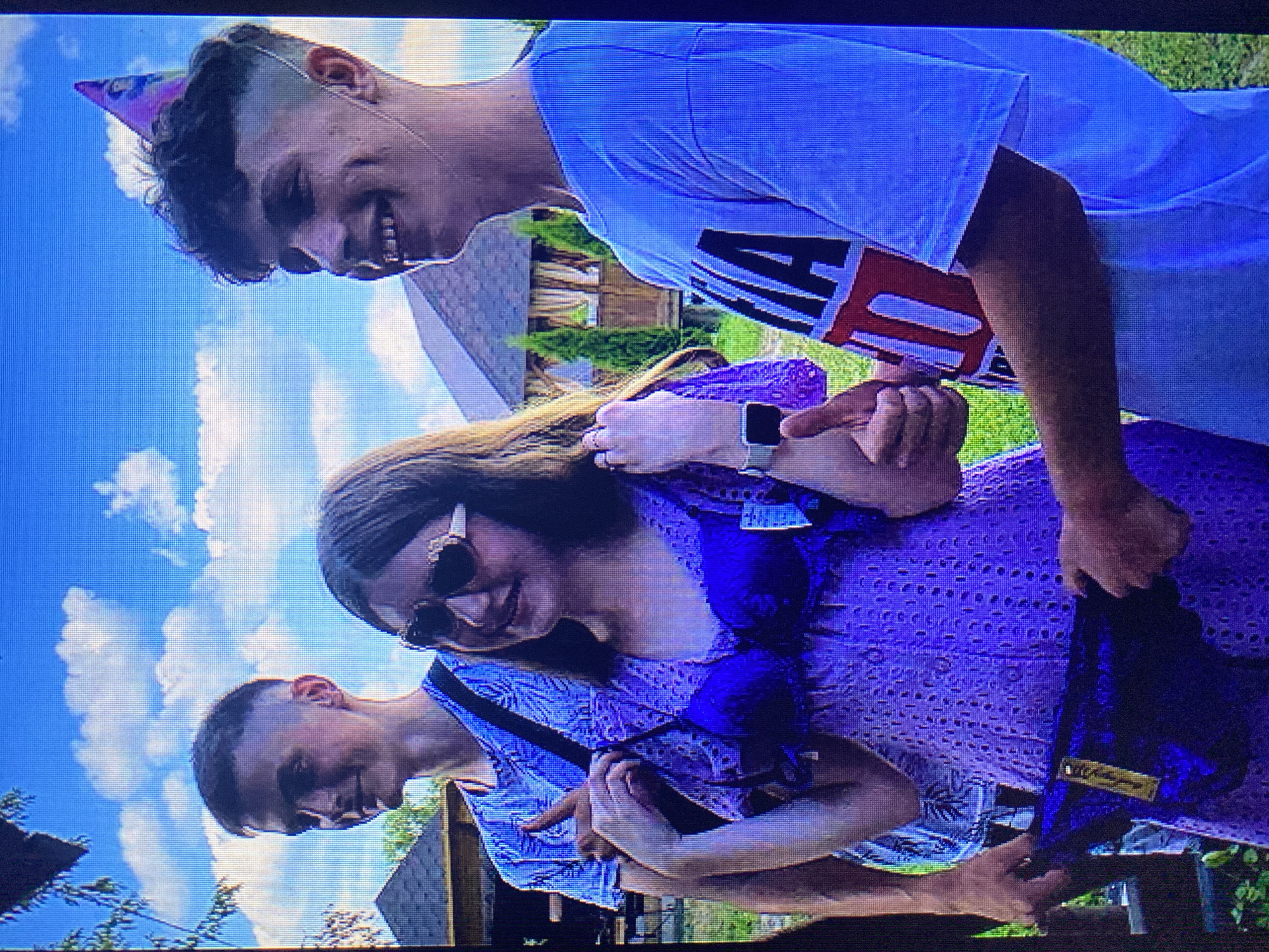Three young people are posing for a photo outdoors. On the left, a young man wearing a blue t-shirt with a palm leaf pattern and a black strap across his chest is giving a thumbs-up gesture. In the center, a young woman wearing heart-shaped sunglasses and a purple lace dress is holding up a matching purple bra and underwear set, with tags still attached. On the right, a young man wearing a white t-shirt with red and black lettering that appears to read "MA 10" is smiling and wearing a purple party hat. The background consists of a blue sky with white clouds, green trees, and a building with a thatched roof. The scene appears to be in a park or garden setting, possibly in the city of Poznan, Poland.