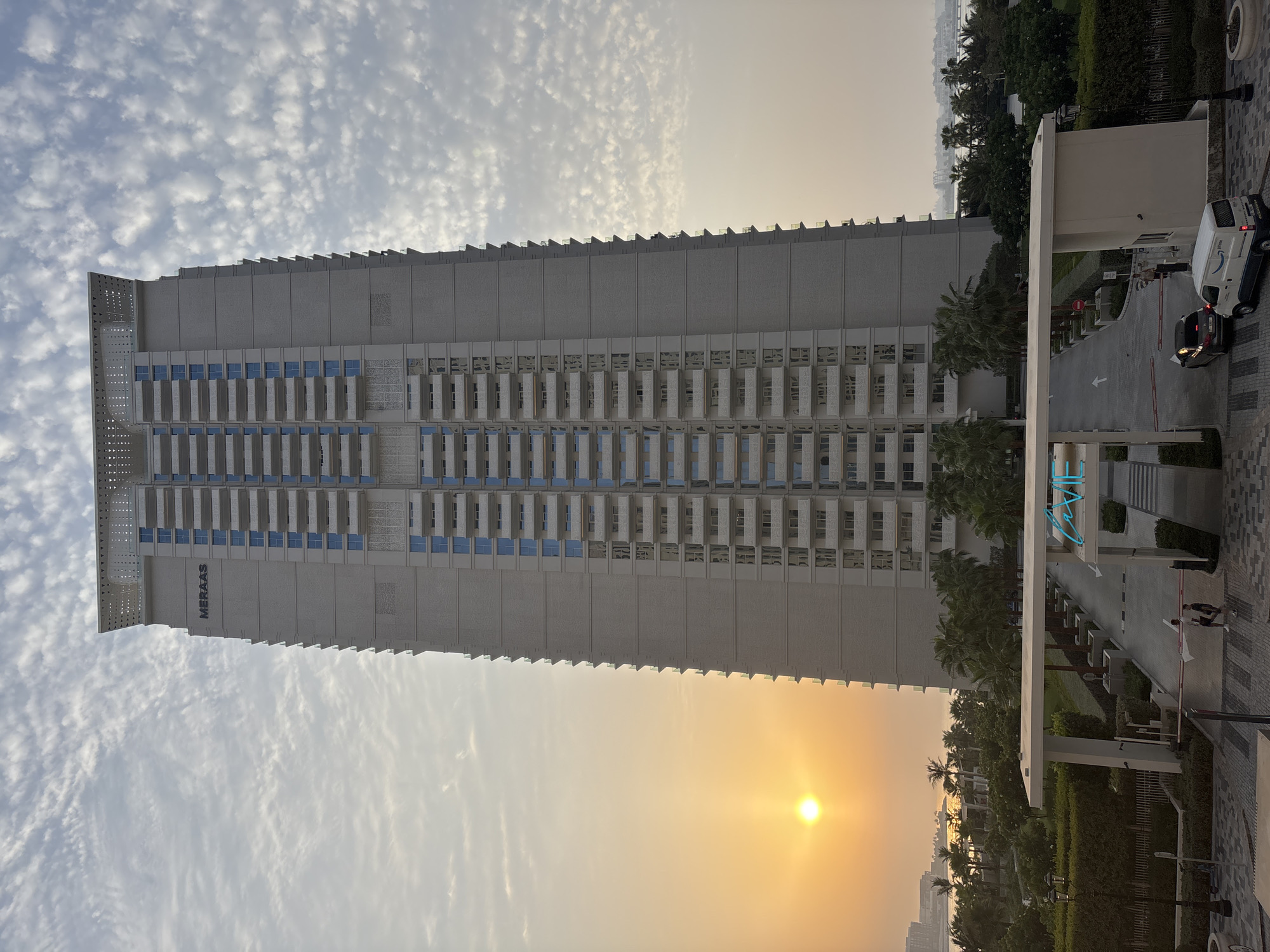 This image depicts a modern, tall, light-colored residential building with numerous balconies, featuring the "MERAAS" logo near the top. The building is situated in Dubai, United Arab Emirates, as indicated by the architectural style and the presence of palm trees and a coastal view in the background, which appears to be the Dubai coastline.
The scene captures a warm sunset with the sun low on the horizon, casting a golden glow on the sky and the left side of the building. The sky is partly cloudy with wispy clouds. In the foreground, a white gateway with illuminated blue lettering that reads "VIE" marks an entrance. A security booth is visible to the right of the gateway. Two individuals are walking towards the gateway, and two vehicles, a dark car and a white van, are entering the premises. Lush green landscaping, including trees and hedges, surrounds the entrance and extends towards the building. The overall atmosphere suggests a residential or mixed-use development in an urban coastal setting.