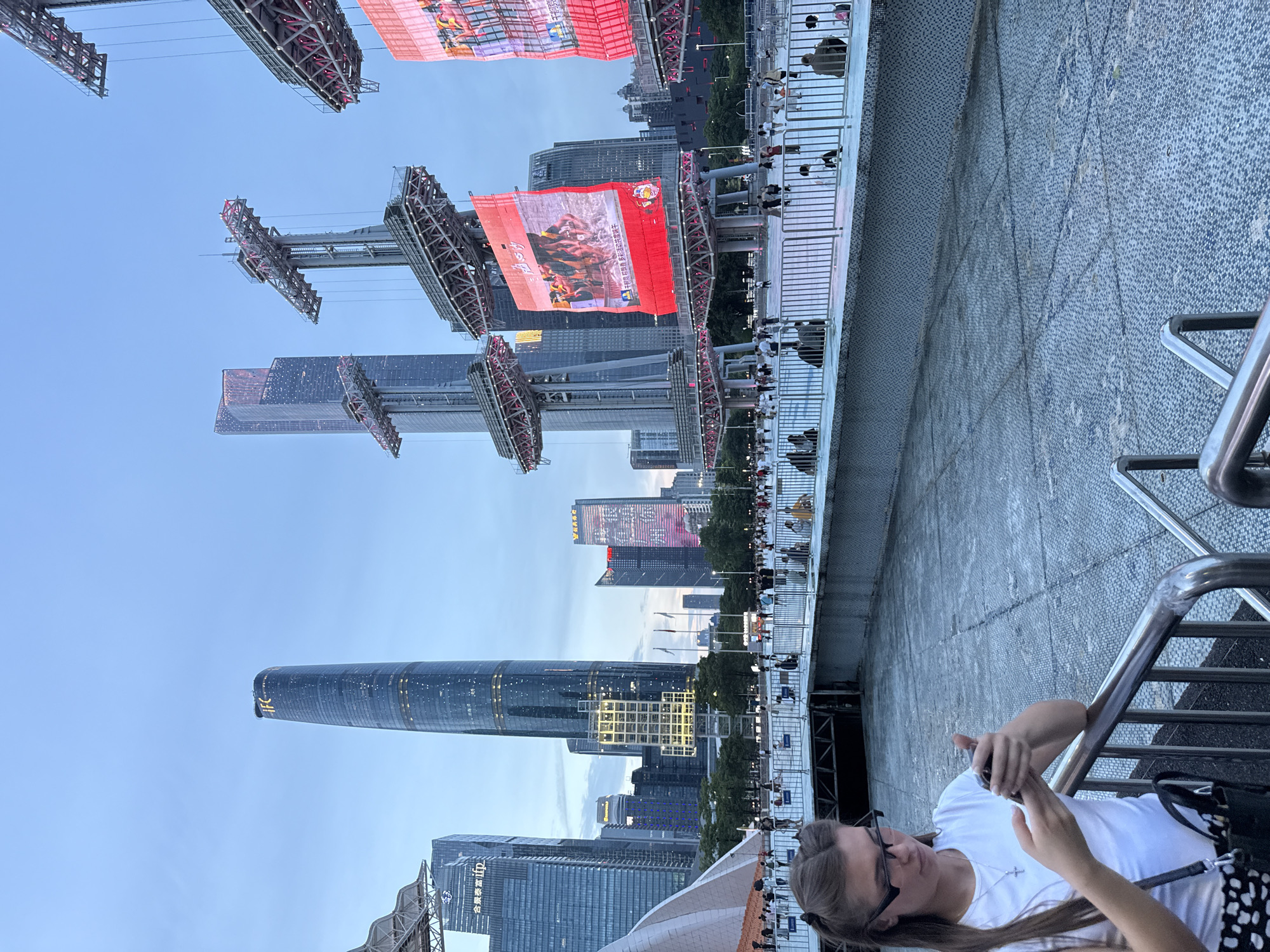 The image depicts an outdoor scene in Tianhe District, China, likely during the early evening hours, as indicated by the twilight sky and illuminated skyscrapers. In the foreground, a young woman with blonde hair pulled back and wearing glasses is looking down at her smartphone, holding it in her hands. She is wearing a white t-shirt and a black and white polka dot purse is slung over her shoulder. A metal railing is in front of her.
In the middle ground, a large crowd of people is gathered, separated from the foreground by a raised walkway with metal railings. The people are milling about, standing, and some are seated. Beyond the crowd, a massive, illuminated LED screen is displaying vibrant imagery. There are also several large, modern skyscrapers with complex lighting visible in the background, including the IFC tower which is prominently lit with gold. The overall environment suggests a public event or gathering in a busy urban setting.