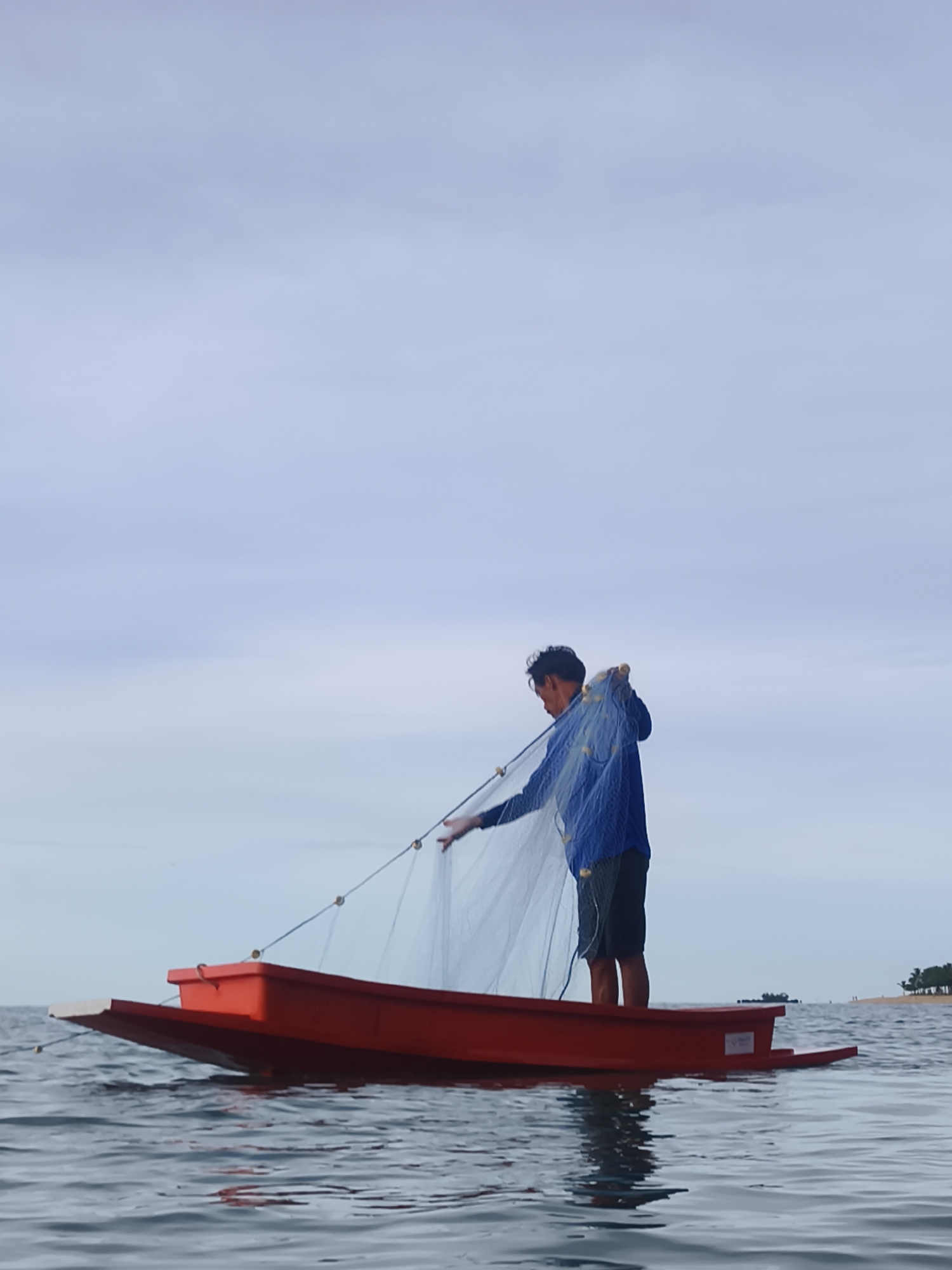A man stands in a small, bright red boat on the water, casting a fishing net. The man is wearing a blue long-sleeved shirt and dark shorts. The boat is a simple, shallow vessel with a light-colored front and red hull. The water is calm, with gentle ripples reflecting the overcast sky. The sky is a pale blue with wispy clouds, suggesting it is either early morning or late afternoon. In the distance, a tree-lined shore is visible, with a few palm trees dotting the horizon. The scene evokes a sense of quiet activity and traditional fishing practices. There is no visible text in the image.