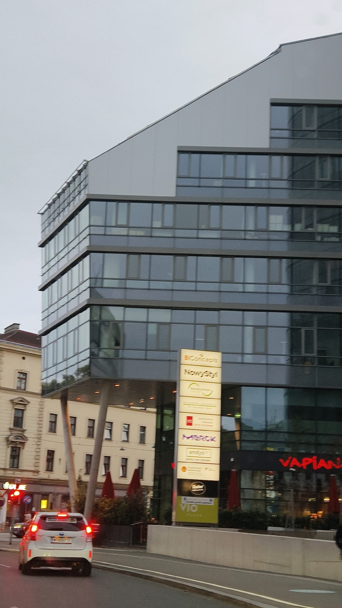 This is a photograph taken from a street level, likely from a moving vehicle, in Vienna, Austria. The primary subject is a modern, multi-story glass and metal building with a distinctive angular design. The sky is overcast and gray.
On the right side of the building, a tall, vertical sign displays multiple logos and company names. The visible names include "BiConcepts," "NowyStyl," "MERCK," "andys," "Tischler," and "VIO." Further to the right, a red neon sign reads "VAPIANO."
To the left of the modern building, an older, cream-colored building with traditional architecture is visible, featuring several windows and architectural details.
In the foreground, a white car with its brake lights illuminated is driving on the street. The license plate is partially visible and appears to have Austrian characters. Traffic lights are visible in the background on the left, with one red light illuminated. The street is paved and shows lane markings.
The environment appears to be urban, with streetlights and mature trees visible, some with red umbrellas protruding from them in front of the Vapiano restaurant. The overall scene suggests a typical city street in the late afternoon or early evening, with overcast weather.