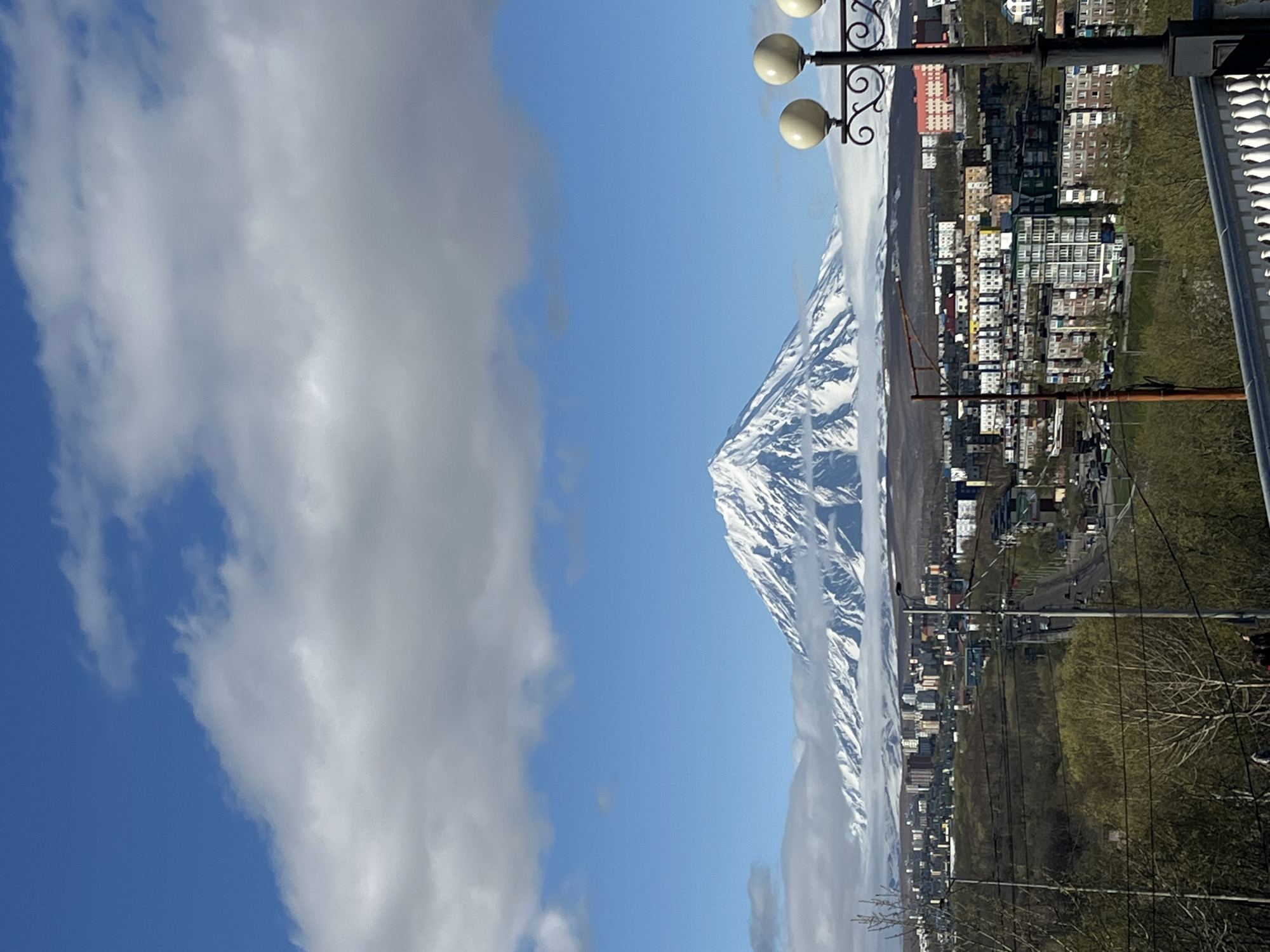 The image captures a view in Petropavlovsk-Kamchatsky, Russia. A snow-covered mountain range dominates the background. A cluster of buildings stretches along the base of the mountain. Electrical wires crisscross the landscape in front of the buildings. A park or forested area borders the buildings. The left side of the image features a blue sky partially obscured by gray and white clouds. A decorative lamppost stands in the upper right corner. A white ledge, likely a railing, is visible along the top right edge.