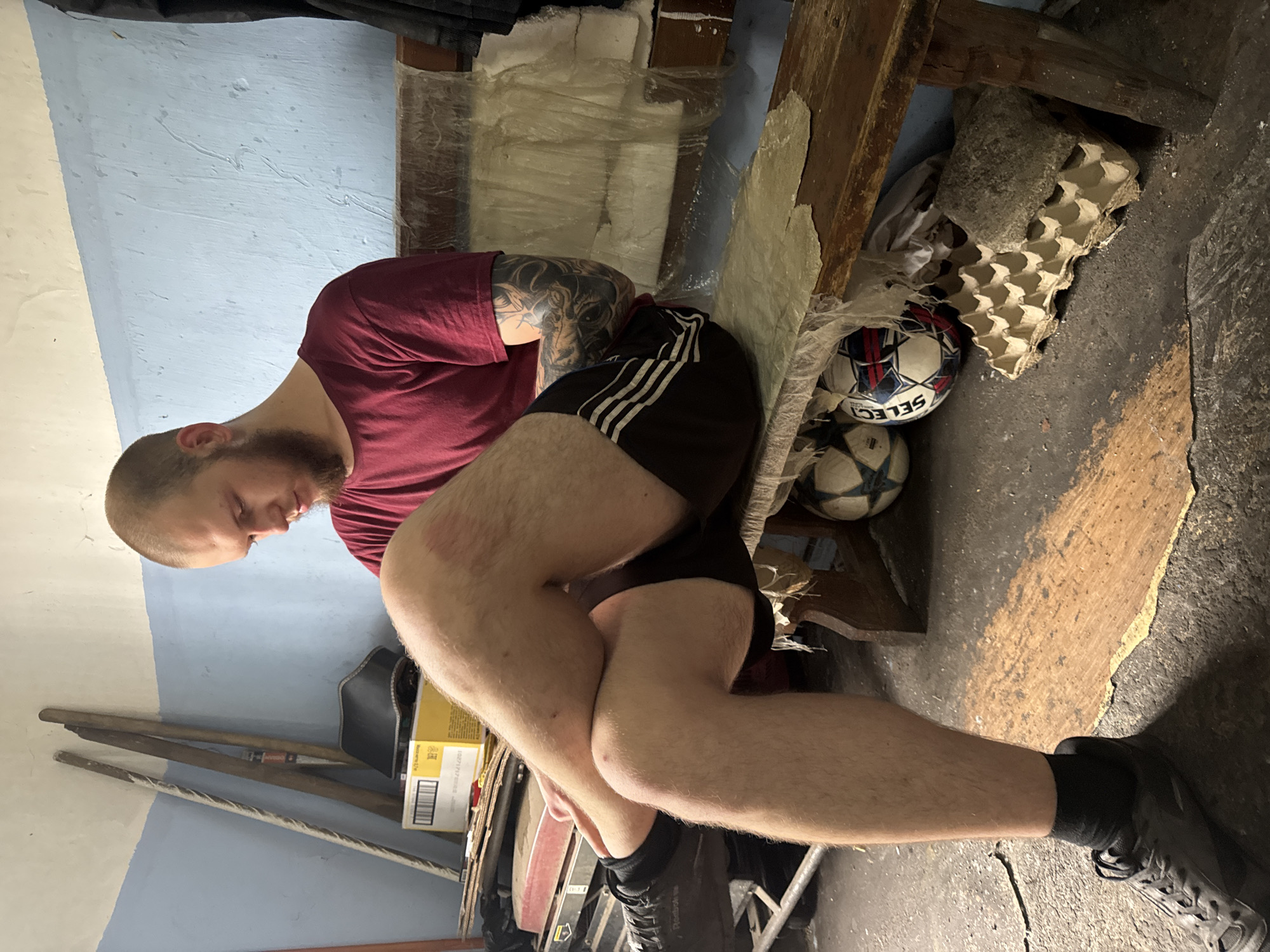 An overhead, medium shot captures a young Caucasian man with a shaved head and a beard, seated on a wooden bench. He is wearing a maroon t-shirt and dark shorts, with his legs crossed and arms resting on his knees. The background reveals a light blue, textured wall, with various objects placed against it, including two wooden poles, a black leather object, and a yellow cardboard box. Below the bench, two soccer balls are visible, along with an egg carton filled with a gray substance. The floor is covered with dirt and debris. The lighting is dim, suggesting an indoor setting.