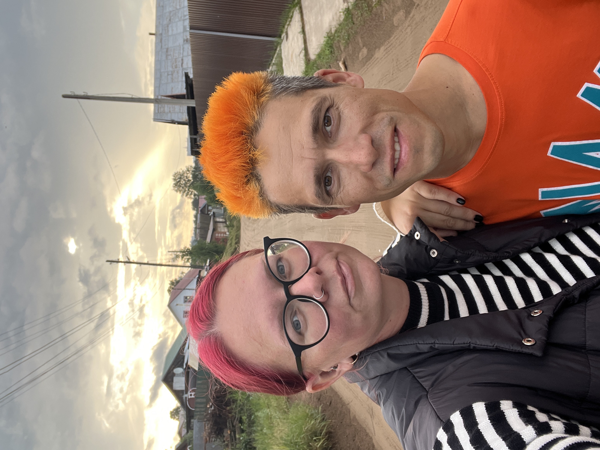 A selfie taken outdoors in Chaikovsky, Russia, features a man and a woman standing on a dirt road. The woman, on the left, has bright pink hair and wears black-rimmed glasses, a nose ring, and hoop earrings. She is dressed in a black puffer vest over a black and white striped sweater. The man, on the right, has spiky orange hair with gray roots and is wearing an orange t-shirt with a teal and white graphic design. His right hand rests on the woman's shoulder. In the background, several houses with metal roofs, power lines, and a wooden fence line the road. The sky is cloudy with sunlight breaking through.