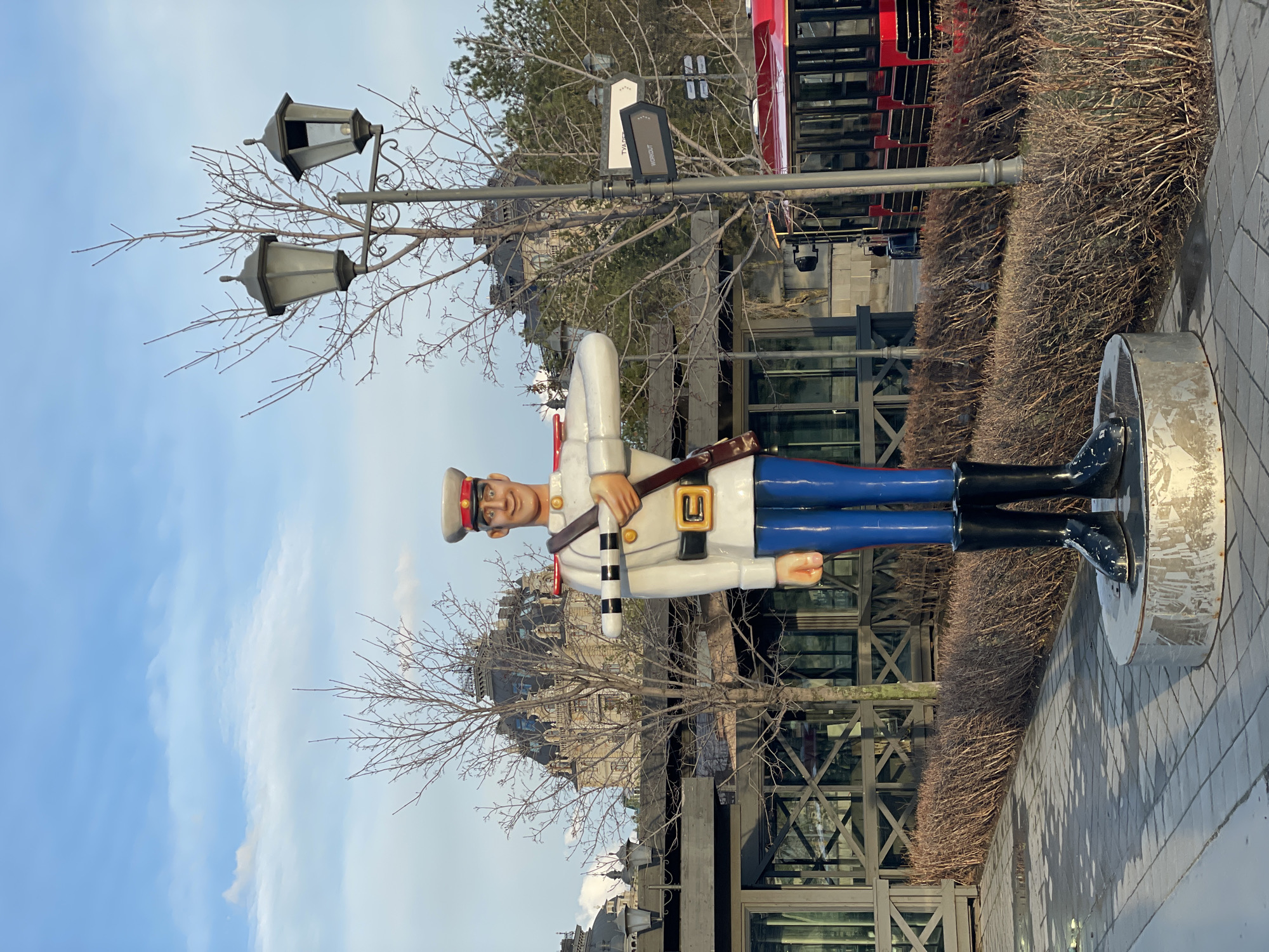 A vibrant vertical image captures a large, colorful statue of a soldier in an urban outdoor setting, likely in Kazan, Russia. The statue, reminiscent of a toy soldier or nutcracker, stands prominently in the foreground, facing the viewer. It wears a white jacket with gold accents, blue trousers, black boots, and a grey cap with a red band, holding a black and white striped baton. Its face is depicted with a friendly, cartoonish smile.
To the left of the statue, a decorative street lamp pole features two multi-lantern fixtures. Bare tree branches, characteristic of late autumn or winter, frame parts of the sky and intermingle with the urban elements. In the background, a mix of architectural styles is visible, including buildings with reflective windows and others displaying ornate detailing. A bright red double-decker bus is partially seen on the right side of the frame, suggesting a tourist or transport route.
The ground is a paved sidewalk. The sky above is a clear blue with scattered white clouds, indicating daytime, possibly late afternoon due to the soft light. Faint text "URAL" and "WORKOUT" are visible on signs attached to a pole in the background, hinting at local area names or activities. The overall scene blends whimsical art with everyday urban life.