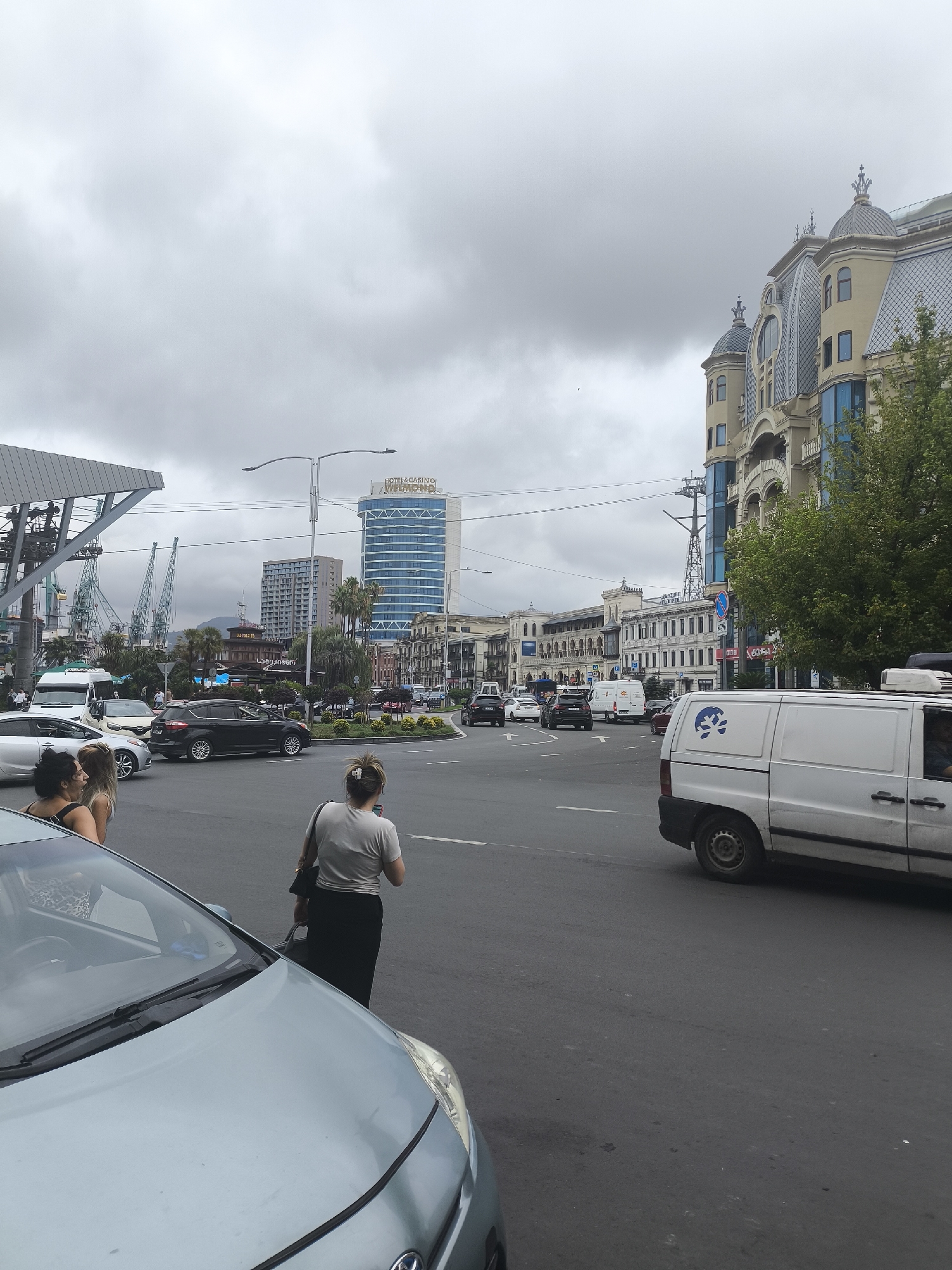 The image shows a street scene in Batumi, Georgia, with a focus on an intersection. The sky is overcast with heavy clouds. In the foreground, to the left, is the hood and windshield of a light blue car. To the right of the blue car, a woman wearing a grey t-shirt and a long black skirt is walking away from the camera, holding a dark bag. Behind her, to the left, three women are standing on the sidewalk near a silver car.
The intersection is busy with traffic. A white van with a blue logo on its side is visible in the right foreground, driving away from the camera. Other vehicles, including a black SUV and several cars, are seen in the intersection and on the street. In the background, there are various buildings, including a tall blue building with "HOTEL & CASINO WEEKEND" signage, and older ornate buildings with decorative facades and mansard roofs. Cranes and industrial structures are visible in the distance to the left, suggesting proximity to a port. Trees and palm trees are scattered throughout the scene, indicating a temperate climate. The road surface is asphalt, and lane markings are visible.
