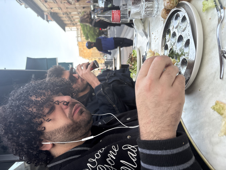 The image captures an outdoor cafe scene on a street in Paris, France, during daylight hours. Two males are seated at a white table. The primary subject in the foreground, a male with dark, curly hair and a beard, wears a black jacket with white embroidery and striped cuffs. He has white wired earbuds inserted and is looking down, his right hand holding a piece of bread over a silver escargot plate that contains green residue in its wells. Empty escargot shells and additional bread pieces are scattered on the table around the plate. To his right, a second male, partially visible, wears sunglasses and a dark denim jacket while drinking from a glass containing a dark beverage. A plate of green salad is positioned in front of this individual. Also on the table are a red-labeled Coca-Cola bottle, a clear water glass, and various cutlery. The background reveals a street with a child in a blue and black jacket and a dark helmet walking away from the camera, along with other distant pedestrians. The street is lined with multi-story buildings characteristic of Parisian architecture, featuring light-colored facades and dormer windows.