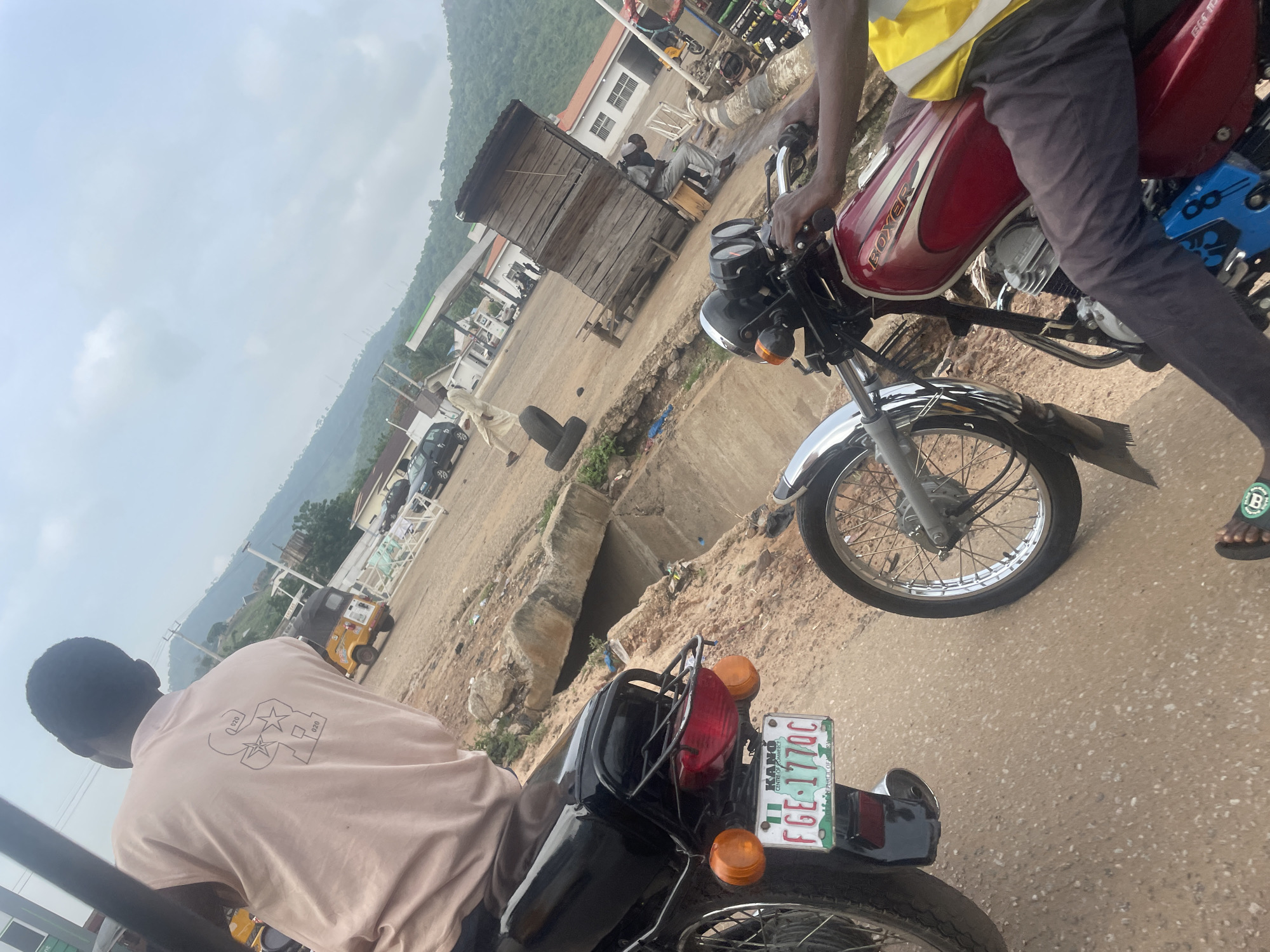 This image captures a street scene in Lokoja, Nigeria, with a focus on two motorcycles and their riders. In the foreground, a person wearing a light-colored t-shirt is seated on a black motorcycle, facing away from the camera. The motorcycle has a green and white license plate reading "KANO FGE 177QC". To the right, another person, wearing a yellow vest and dark pants, is on a red motorcycle, also facing away from the camera. The red motorcycle has "BOXER" visible on its fuel tank.
The environment suggests a somewhat dusty and utilitarian area. A concrete drainage channel runs along the right side of the road, with some debris visible. In the background, there is a fuel station with pumps and a small wooden kiosk. Hills covered in vegetation rise behind the structures. Several other vehicles, including a yellow auto-rickshaw and a black car, are present in the mid-ground. The sky is overcast. The overall impression is of a typical roadside commercial area in Lokoja, likely a place where people commute and conduct business.