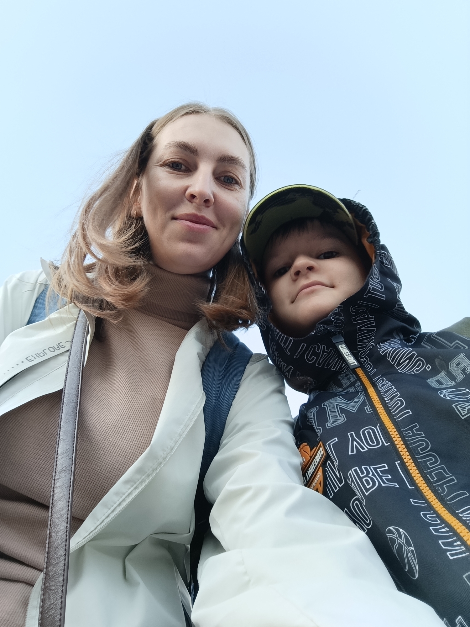A low-angle outdoor portrait features a woman and a child looking down at the camera, likely a selfie. The background is a clear, light blue sky, indicating daytime and good weather.
The woman, on the left, has shoulder-length, light brown hair and appears to be smiling slightly. She is wearing a light-colored, possibly off-white, jacket over a tan ribbed turtleneck. A dark strap from a bag or purse is visible over her left shoulder. Part of the word "EMBROIDERY" is visible in reverse text on her jacket.
On the right, a child, likely a boy, wears a camouflage-patterned baseball cap under the hood of a dark jacket. The jacket has a vibrant yellow zipper and is patterned with various white letters and shapes, including a basketball graphic. Visible text includes fragments like "M", "JUM", "AO", and "BE", some of which are inverted. The zipper pull has the text "20E3CHT". The child has a neutral expression, looking directly at the camera.
The overall impression is a casual, happy moment shared between the two subjects. The scene is set outdoors in Bolshoye Savino, Russia.