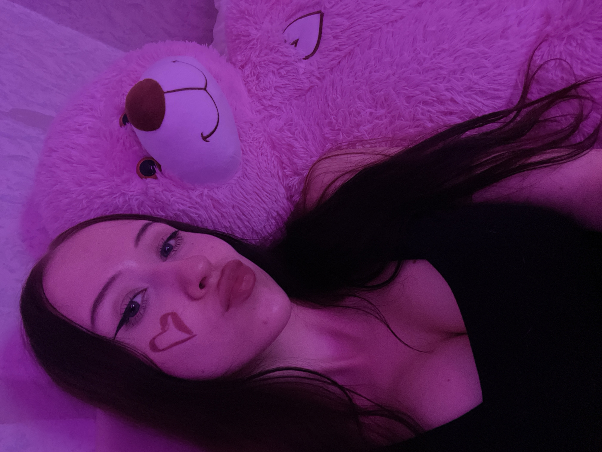 The image is a close-up, high-angle selfie of a young woman posing with a large pink teddy bear. The lighting is a vibrant pink, creating a somewhat surreal and intimate atmosphere. The woman has long, dark hair and is wearing a black top. She has winged eyeliner and a red heart drawn on her left cheek. Her lips are pursed, as if for a kiss. The teddy bear, to her right, is plush and appears to be quite large, filling a significant portion of the frame. Its face is visible, with smiling eyes and a stitched nose. The background is a soft, textured surface, possibly a blanket or rug, also illuminated by the pink light. The setting is indoors, and there is no discernible context suggesting a specific location in Votkinsk, Russia.