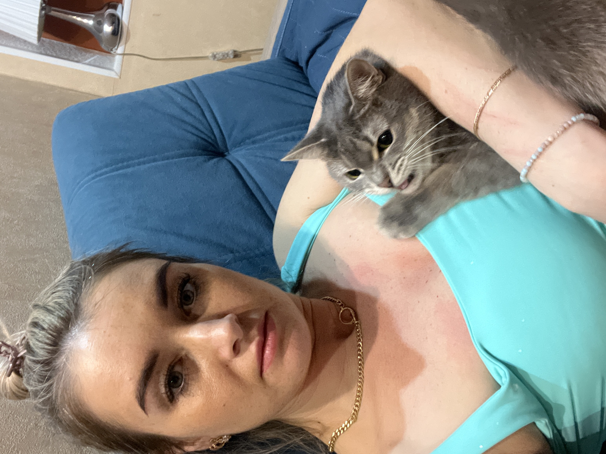 A woman is lying on a blue upholstered piece of furniture, possibly a sofa or daybed. She is wearing a turquoise top and gold-colored jewelry, including a necklace and bracelets. A grey tabby cat is positioned on her arm and chest, with its head near her neck. The setting appears to be indoors, possibly in a home. The background includes a wall and a lamp fixture. There is no visual information available to determine the location as Chaikovsky, Russia.