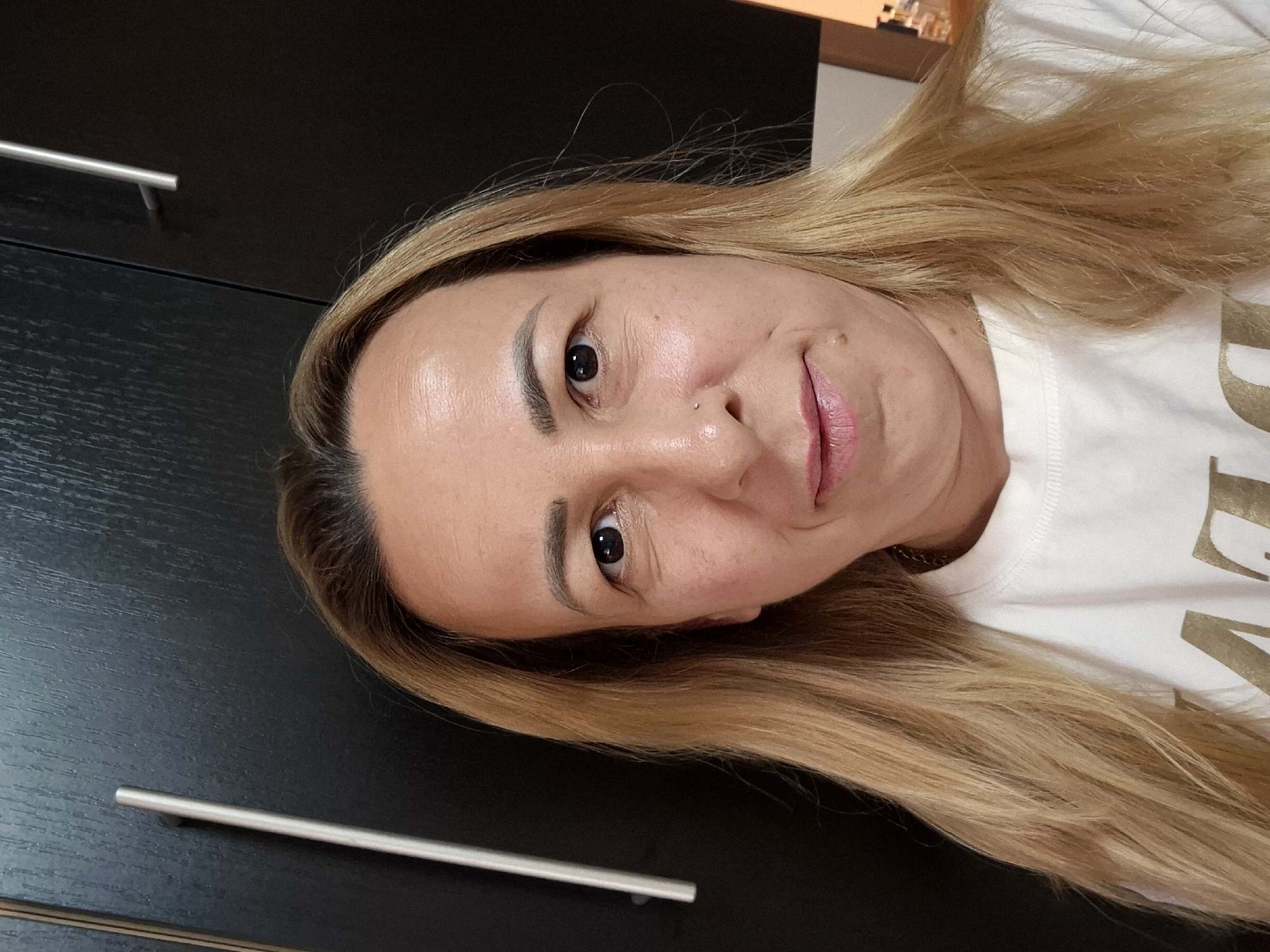 This media file features a close-up, tilted selfie of a woman with blonde hair. She is looking directly at the camera with a neutral expression, her head slightly tilted. Her makeup includes defined eyebrows and lipstick. She has a small nose piercing. Her hair is long and light brown with blonde highlights, falling around her face and shoulders. She is wearing a white t-shirt with gold lettering, though the text is mostly obscured.
The background consists of dark cabinetry with silver handles, suggesting an indoor setting, possibly a bedroom or living area. The lighting appears to be artificial and even, with no clear indication of time of day or weather. The overall impression is a casual, personal photograph.