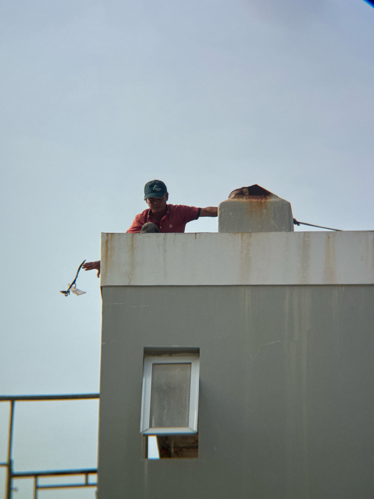 A man wearing a red shirt and a dark baseball cap is on the rooftop of a building, leaning over the edge. He is holding a thin, dark object with white paper attached to its end in his right hand. The man appears to be in his 20s or 30s and is looking down and to his left. In the background, there is a large, weathered concrete water tank with rust stains. The building is painted a light gray color and has a white horizontal band along the top edge. A small, open window is visible on the gray wall of the building. The sky is overcast and a light blue. This scene is observed from a distance, suggesting it might be in a residential or industrial area of Nha Trang, Vietnam.