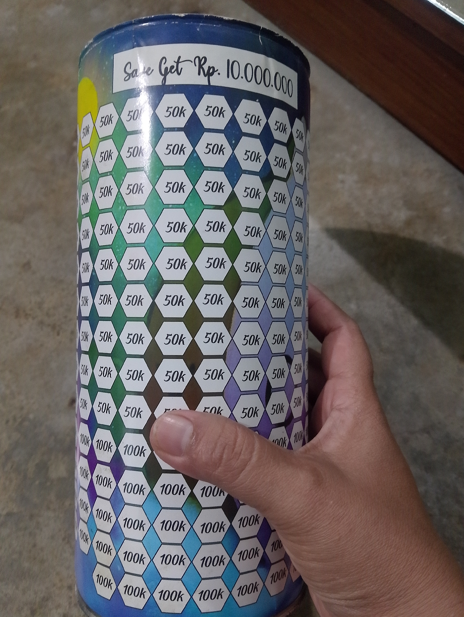 In Serbelawan, Indonesia, a person's hand is holding a cylindrical savings bank. The bank is covered in a hexagonal pattern with text inside each hexagon. The text alternates between "50k" and "100k". A prominent label at the top of the bank reads "Save Get Rp. 10.000.000". The background consists of a floor and a wooden surface.
