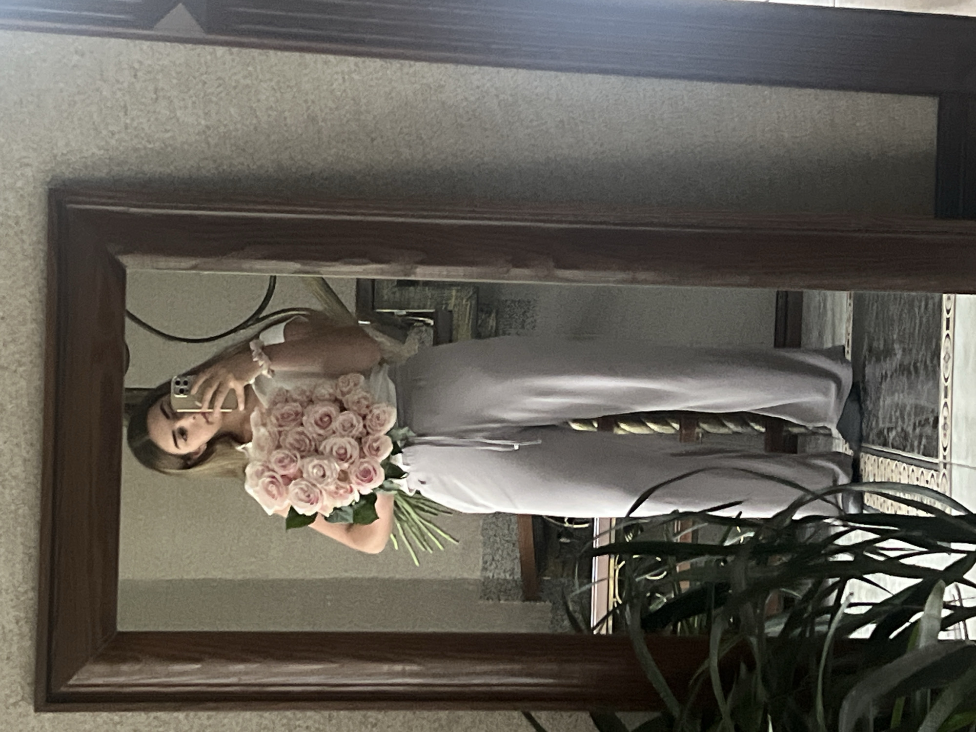 A young woman stands in front of a large, dark wooden framed mirror, taking a selfie with her phone. She is holding a large bouquet of light pink roses. She is wearing a white t-shirt and light gray wide-leg pants. Her reflection is visible in the mirror, showing her face and the bouquet she is holding. In the background of the reflection, there are stairs and a textured wall. In the foreground of the image, a large, leafy green plant is visible, partially obscuring the lower part of the woman's body. The floor appears to be tiled. The setting is indoors, likely a home. There is no indication of the city of Ust-Chorna, Ukraine within the image itself.