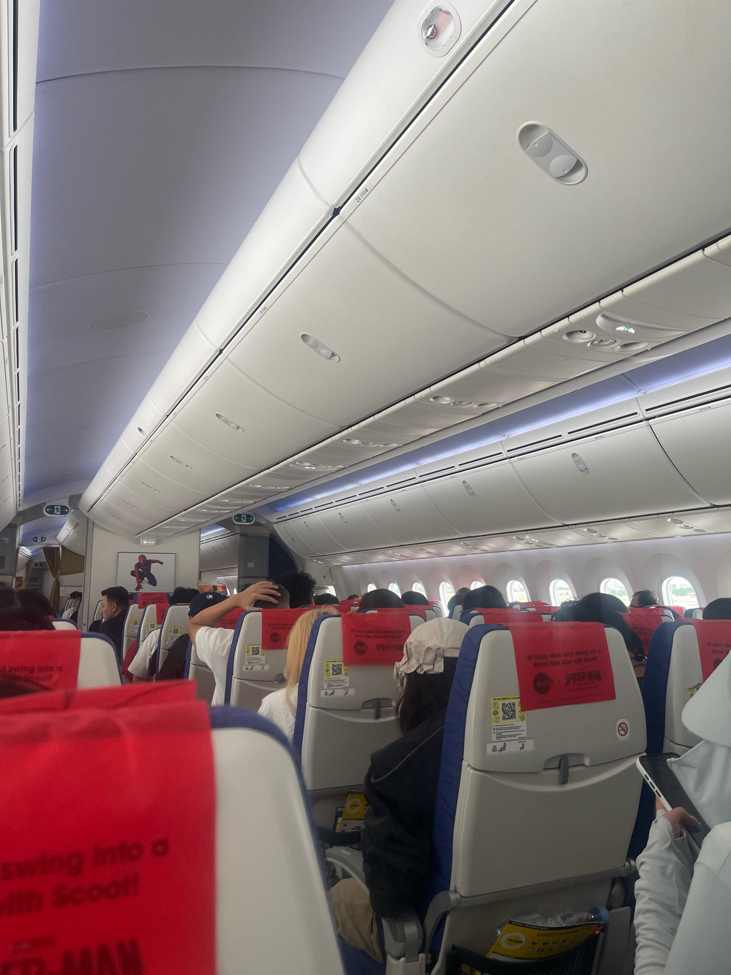 This is a photograph taken from the aisle of an airplane cabin, looking forward. The overhead compartments and lighting fixtures are visible along the ceiling. A framed picture of Spider-Man is mounted on the wall. Passengers are seated in rows of blue and white seats with red headrests. Some passengers are visible from the back and side, with one person in the foreground touching their head. The seats have safety cards and QR codes on the back. The environment appears to be indoors, within an aircraft. The location is specified as Baiyun District, China.
