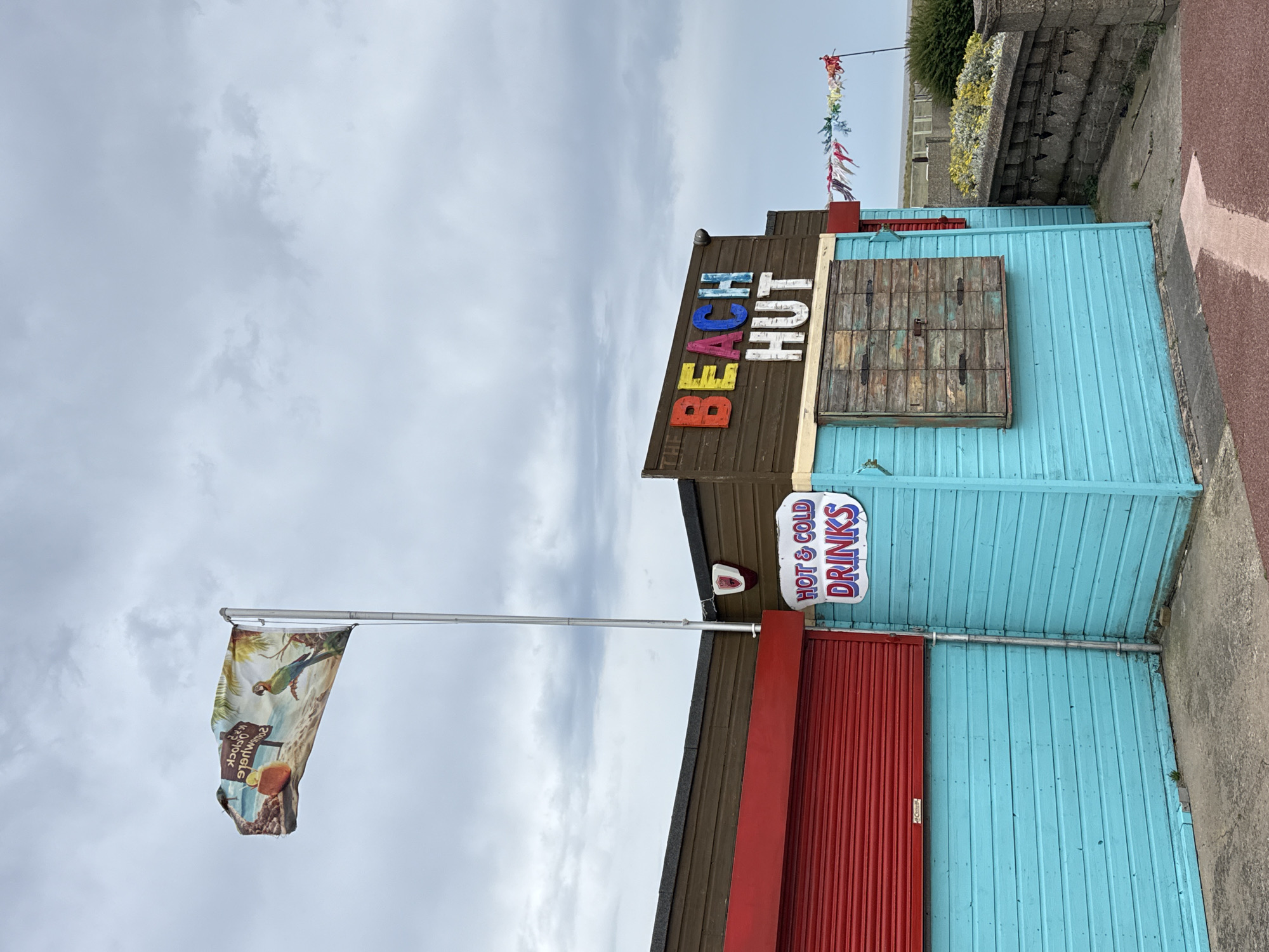 This media file depicts a beach hut in Great Yarmouth, United Kingdom. The hut is painted a vibrant turquoise blue with a brown roof. Above the entrance, the words "BEACH HUT" are displayed in colorful, blocky letters. To the right of the entrance, there is a sign that reads "HOT & COLD DRINKS" in white text on a blue background. A weathered wooden shutter is visible on the upper part of the hut. A red roller shutter door is present on the lower part of the hut.
To the left of the hut, a flag flies from a metal pole. The flag features an image of a parrot on a beach with a signpost. The sky is overcast and cloudy, suggesting a cool or temperate day. In the background, a low wall and some greenery can be seen, and a cobbled path runs along the side of the hut. Colorful bunting is strung above the hut, adding a festive touch. The overall scene conveys a typical seaside establishment, likely a kiosk or small shop catering to beachgoers.