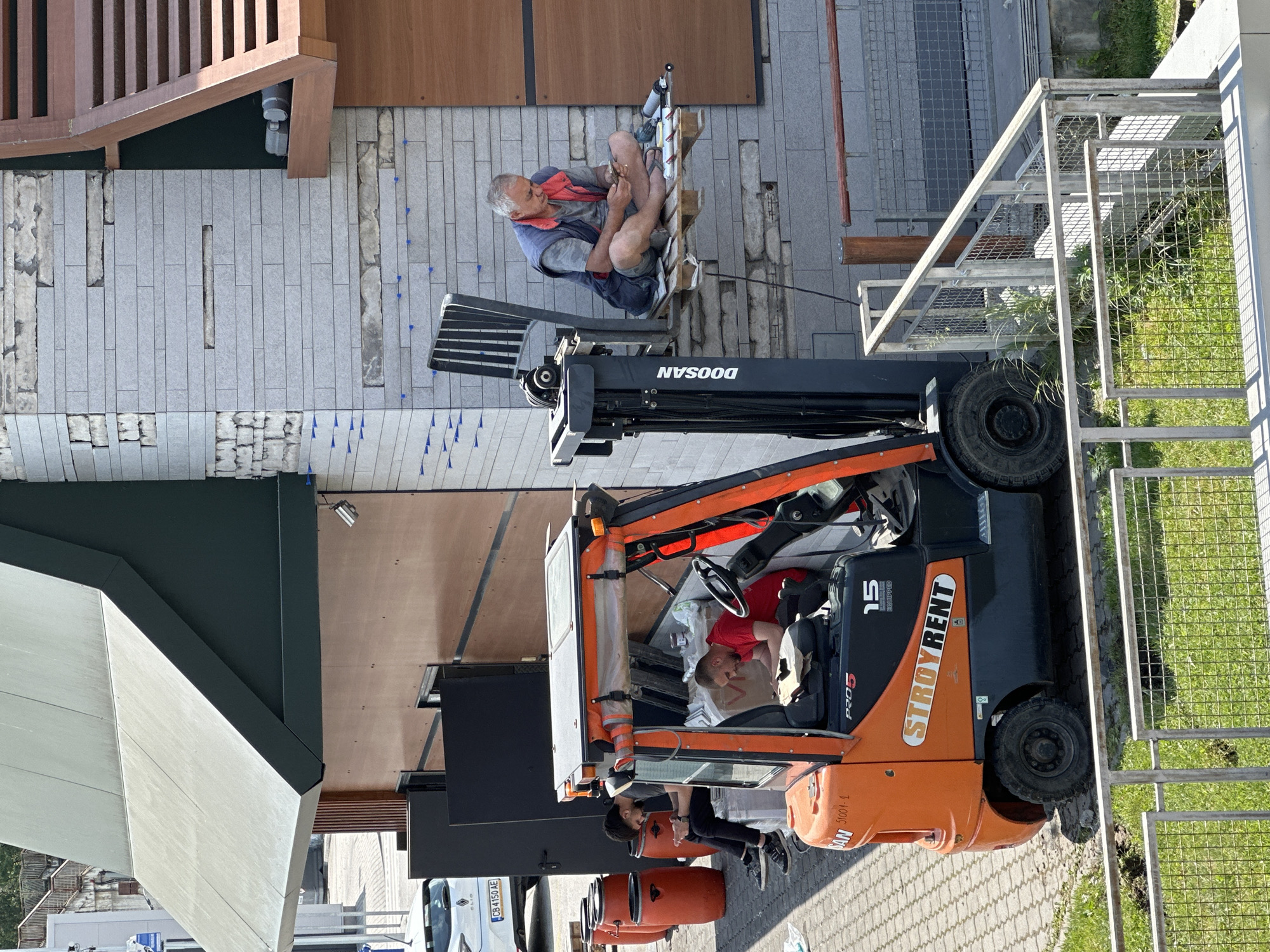 The image, taken at an elevated perspective, shows a construction or renovation site in what appears to be Veliko Tarnovo, Bulgaria. A man is seated on a pallet raised by a Doosan brand forklift, seemingly working on the exterior of a building. He's wearing casual attire: shorts, a t-shirt, a vest, and sandals. The building appears to be undergoing tile work, with tiling partially completed on a facade. Adjacent is a wooden structure, and beyond that, a paved area.
An orange Stroyrent forklift truck has two other individuals. One male in the driver seat and a women is on top of the vehicle.
The environment includes a sidewalk with cobblestone pavement, sections of landscaping with grass and a fence, and a view of surrounding buildings.
The overall scene suggests an ongoing construction or maintenance project in an urban setting.