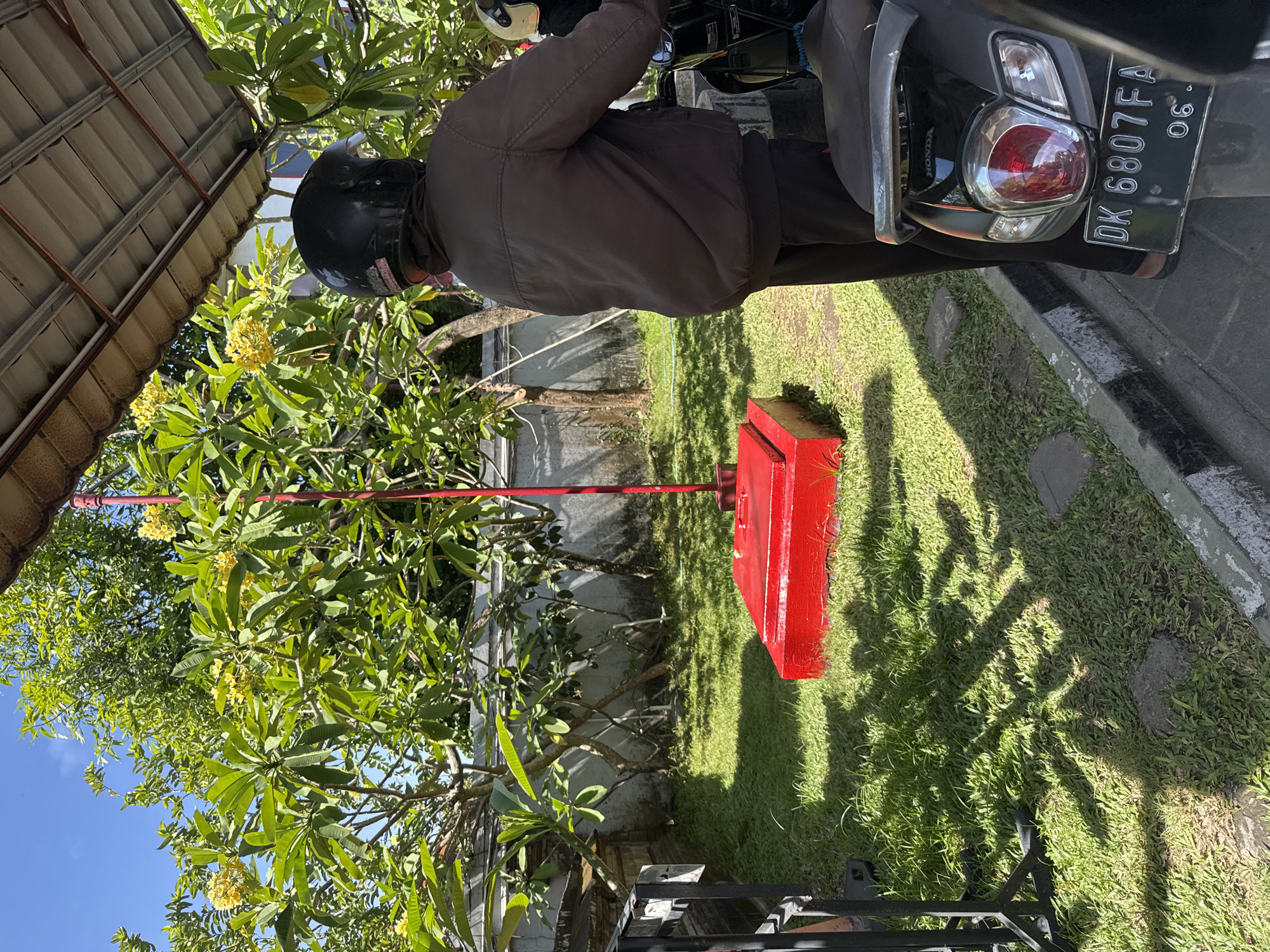 A person wearing a dark brown jacket and a black helmet is standing next to a scooter in Kuta Selatan, Indonesia. The person is facing away from the camera and appears to be observing something to their right. The scooter has a license plate that reads "DK 6807 FA 06". In the background, there is a vibrant green lawn with shadows cast from trees and what appears to be a bright red base with a red pole extending upwards. The environment is lush with green trees and foliage, suggesting a tropical setting. The sky is clear and blue.