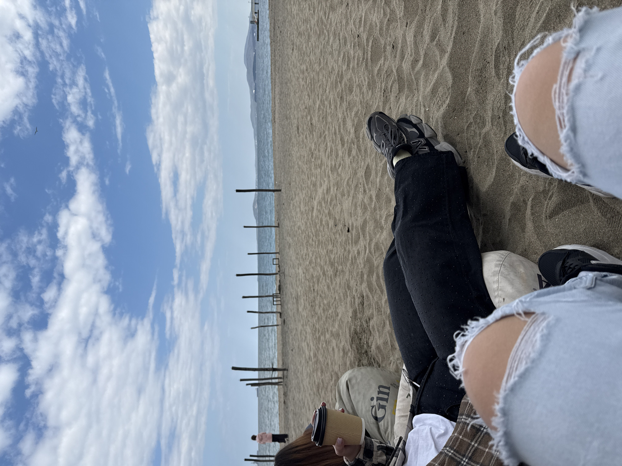 The image captures a beach scene, likely during the day, with a focus on two individuals relaxing. The foreground shows the legs of the people, one wearing black leggings with dark grey sneakers and the other with ripped light blue jeans and black sneakers. They appear to be sitting on the sand on a blanket. In the midground, a person holds a coffee cup. The background reveals the sea and a distant coastline or possibly a hill. There is a line of wooden posts extending into the sea, likely wave breakers. The sky is visible with clouds. Given the information available, it's impossible to confirm the location as Burgas, Bulgaria, without more identifiable landmarks.