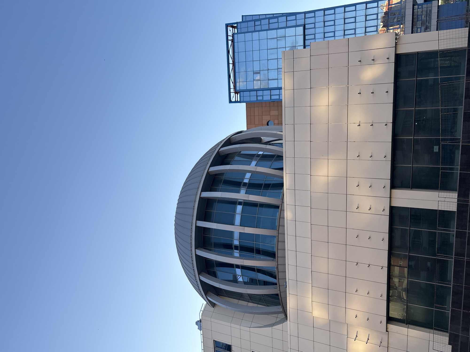 The image shows a modern building complex in Moscow, Russia, under a clear blue sky. The architecture features a domed glass structure adjacent to a white tiled building with a section of dark glass windows. Above, a boxy blue glass structure is visible. The buildings are angled, creating a dynamic composition against the plain sky.