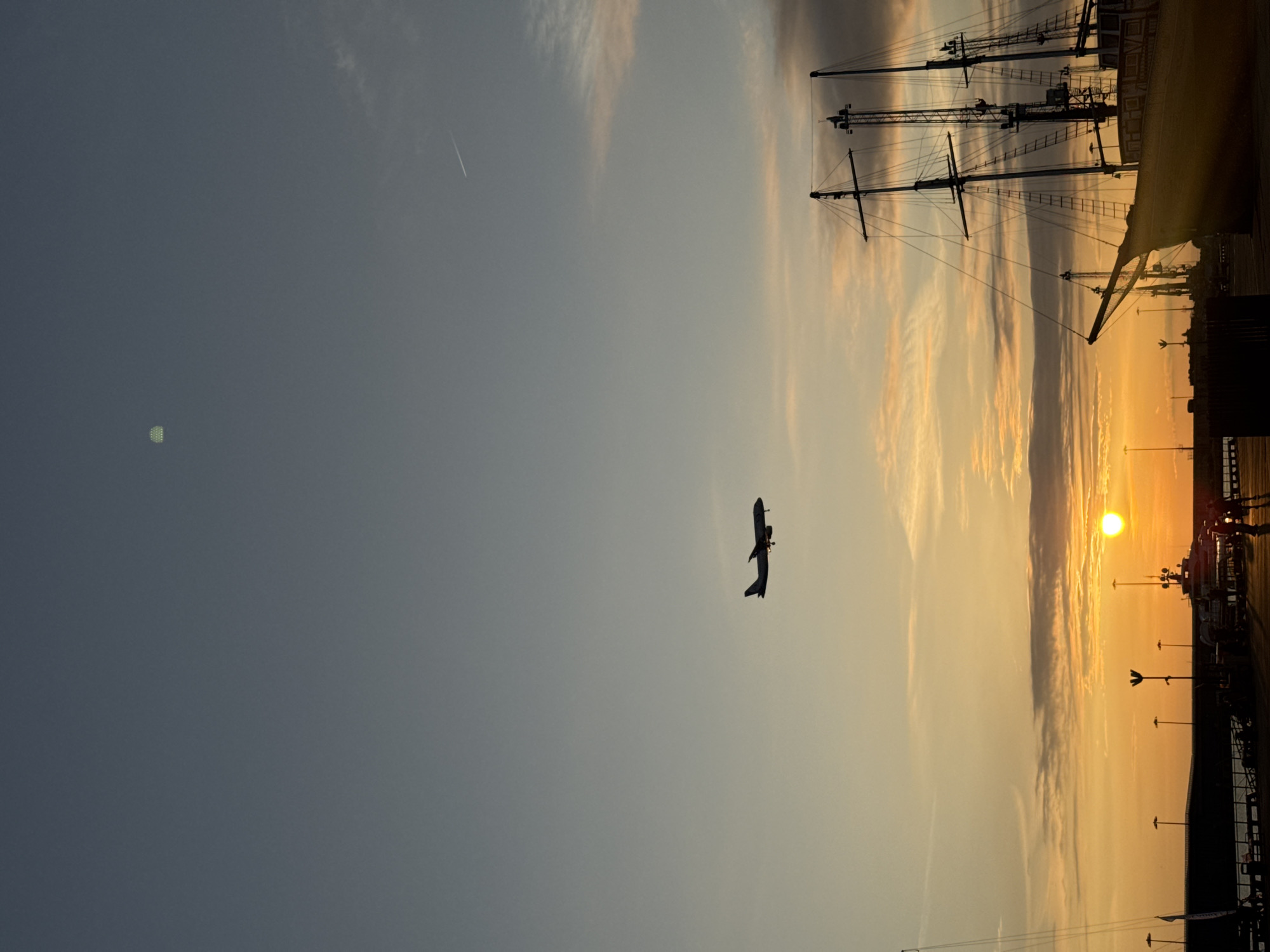 The image presents a sunset scene. In the foreground, there's the silhouette of an airplane against the sky. The background is dominated by the setting sun and a section of the ship. The exact location in the city of Sirius, Russia cannot be determined without more information. There are some unidentified people are walking around the ship.
