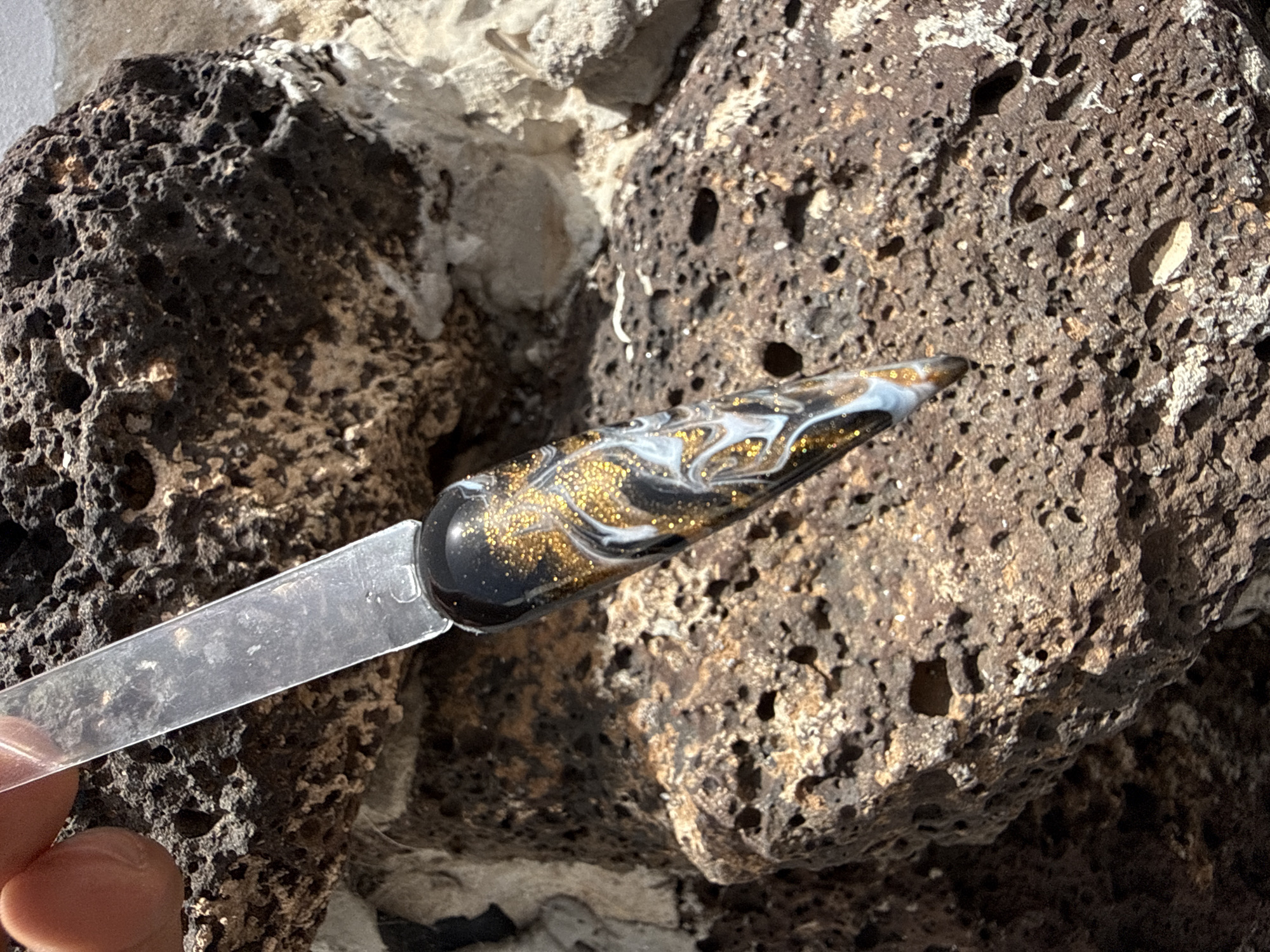 The image shows a nail art sample held against a backdrop of volcanic rock, likely located in or near Yaiza, Spain, given its volcanic landscape. The nail sample features a black base with swirls of white and gold glitter. The rock is porous and dark, indicative of volcanic formation. A finger is visible holding the transparent stick of the nail sample. The lighting suggests a sunny day.