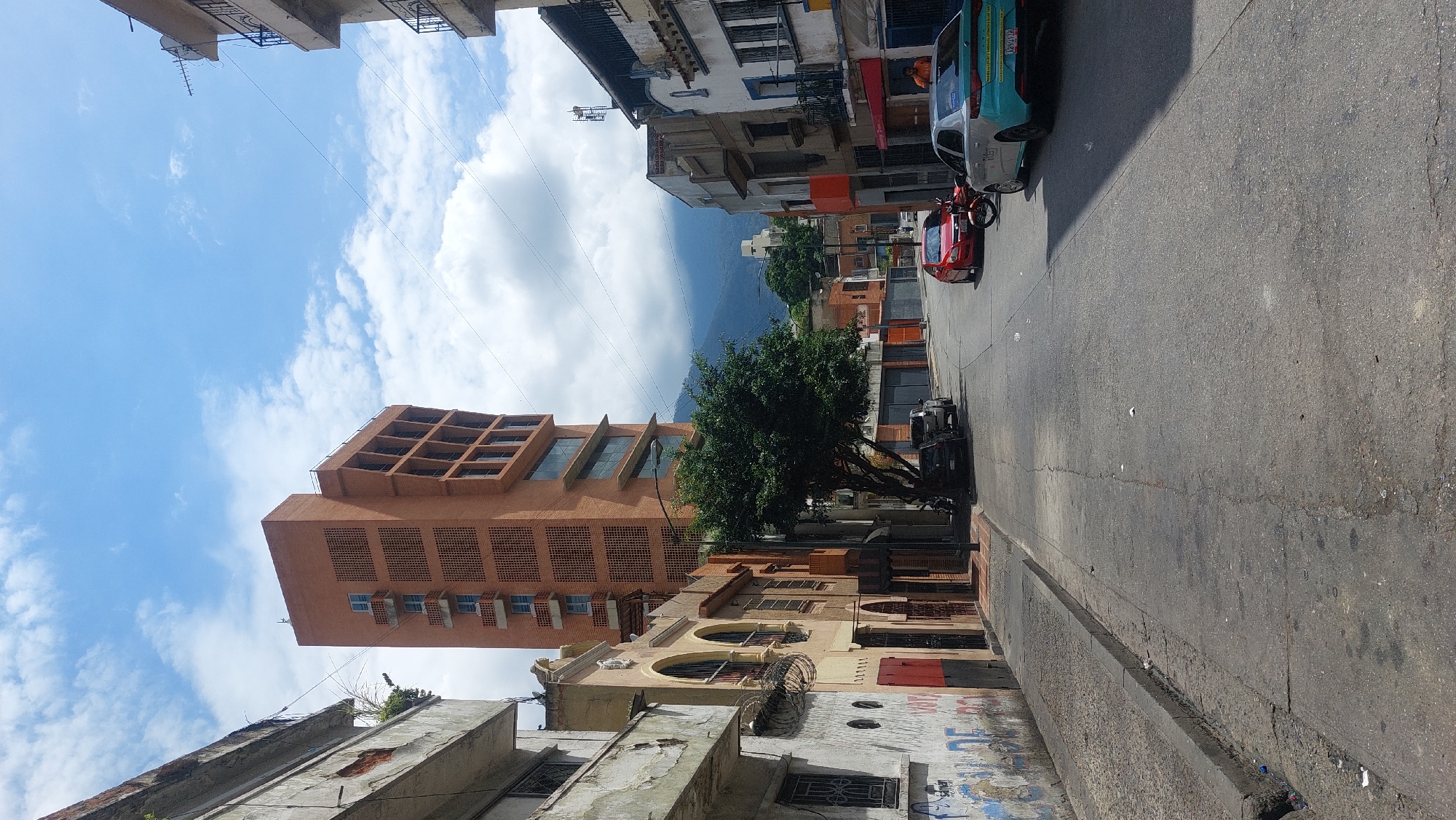 The image captures a street scene in Caracas, Venezuela. The view is taken from street level, looking up the incline of a street. The street's surface shows wear and tear. Buildings of varying heights line the street. On the left, an older, weathered building contrasts with a tall, modern orange-brown building further up the street. The modern building has a grid-like facade. A tree stands along the sidewalk near the buildings. Cars are parked along the right side of the street. In the background, a mountain range is visible, partially obscured by clouds. The sky is blue with scattered white clouds.