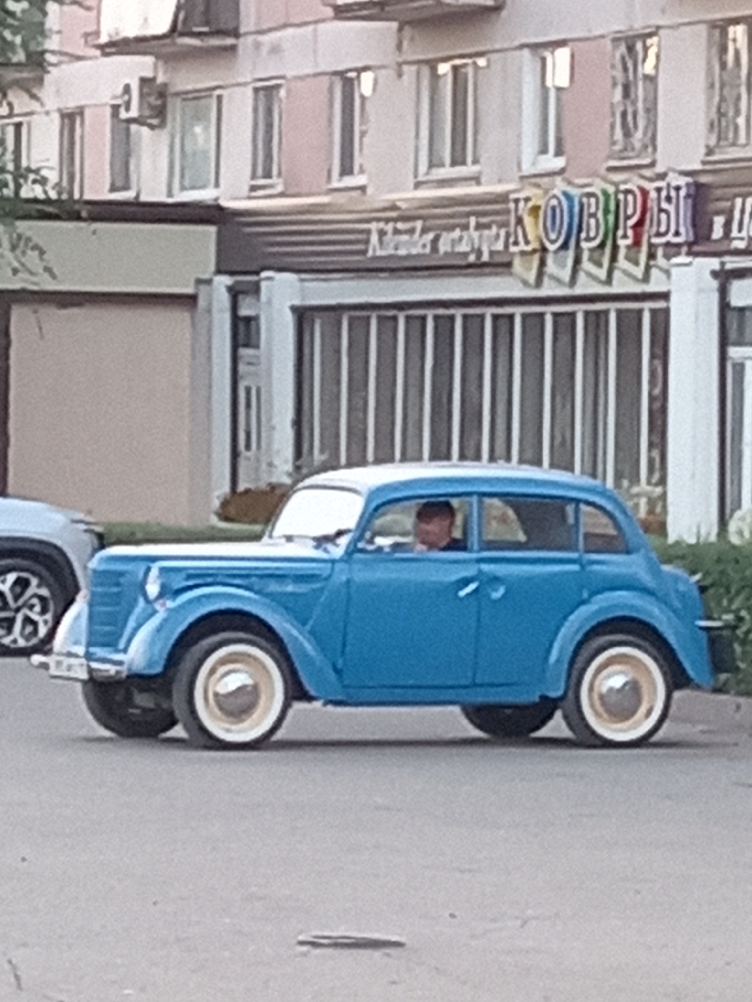 The image shows a blue vintage sedan parked on a street in Rudny, Kazakhstan. The car is the main subject, with a person visible inside, driving. Behind the car, a building with several windows and an air conditioning unit is visible. A sign with colorful letters spelling "КОВРЫ" (which translates to "Carpets") is displayed on the building. The street appears to be paved, and there is some greenery on the right side of the image. The overall scene suggests an urban environment with a touch of retro charm due to the presence of the classic car.