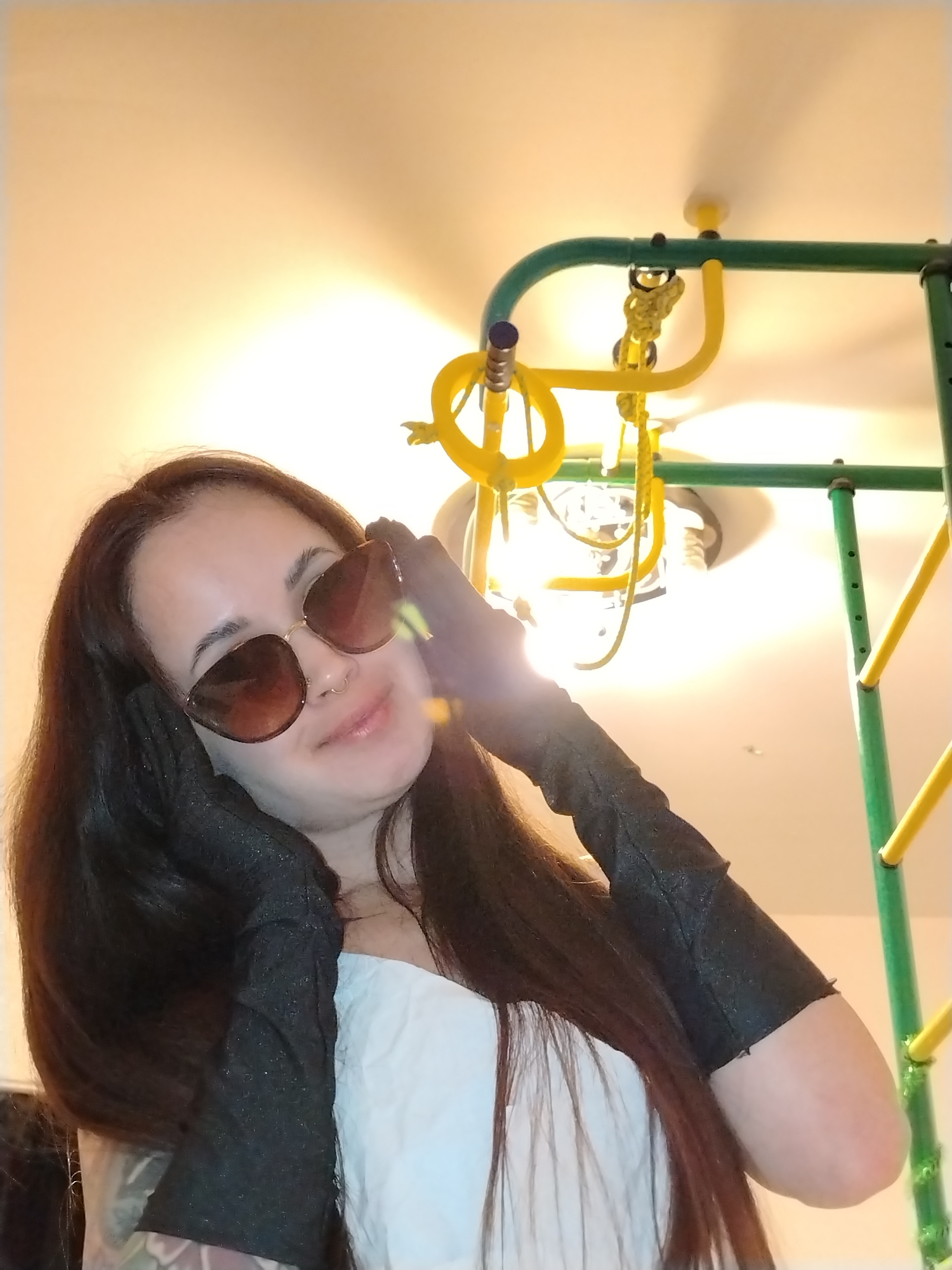 A young woman with long, dark brown hair is depicted in a close-up, upward-angled indoor shot. She wears stylish brown cat-eye sunglasses, black fingerless gloves, and a white off-the-shoulder top. A subtle nose ring is visible on her face. Her gloved hands are positioned near her face, possibly adjusting the sunglasses, and she displays a slight, pleasant smile towards the camera.
The background features a bright, artificial ceiling light that emits a strong glare. Partially visible around and above her are components of a green and yellow indoor play structure, including metal bars, plastic rings, and ropes, suggesting a home environment like a child's room or a playroom. The ceiling is light-colored. The image was captured indoors in Noviy, Russia, under artificial lighting, likely as a casual portrait or selfie. There is no visible text in the image.