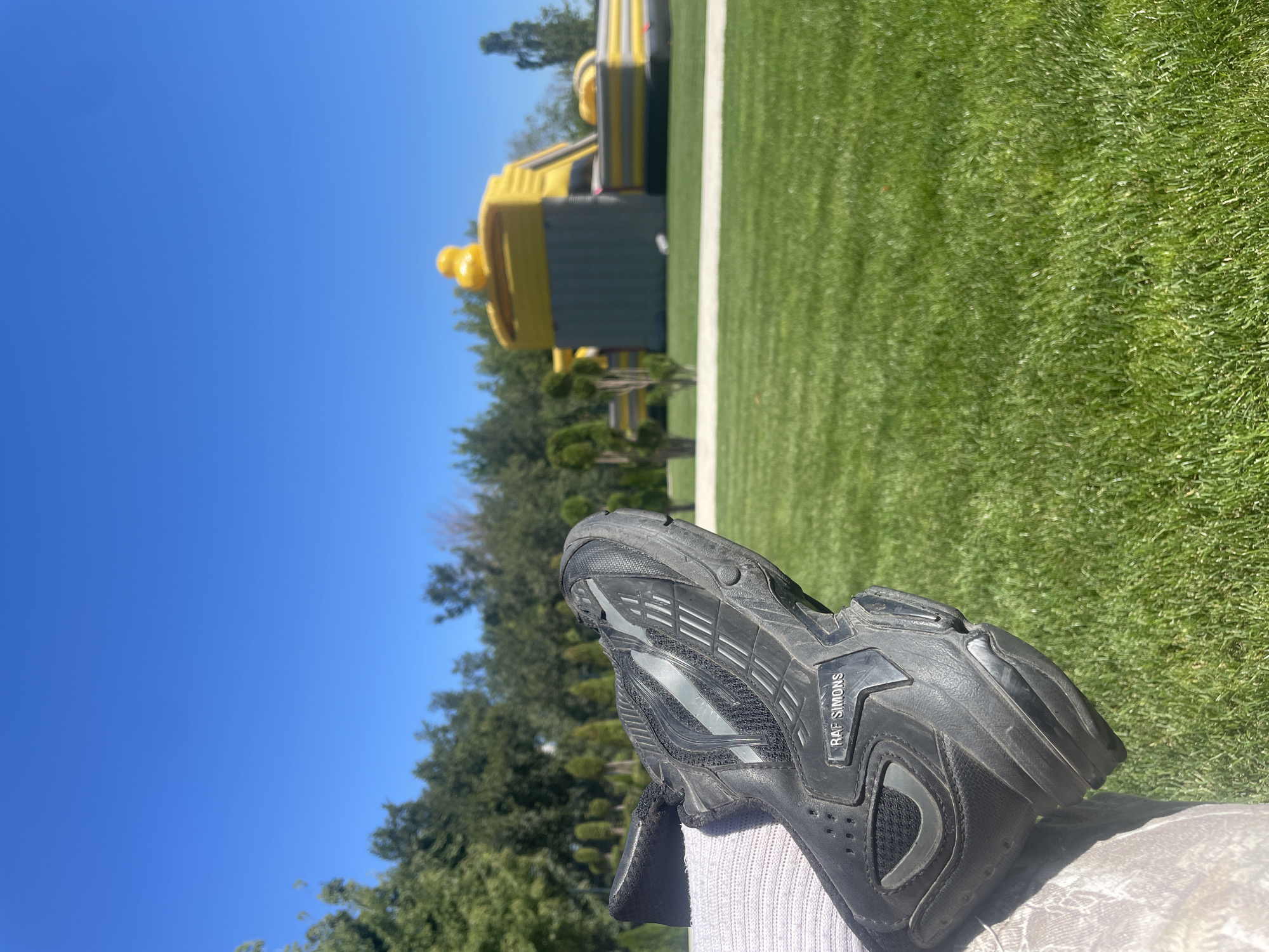 A black Raf Simons sneaker rests on a person's leg, which is clad in a patterned camouflage fabric and a grey sock. The sneaker is positioned in the foreground, angled towards the right side of the frame. Behind the sneaker, a vibrant green lawn stretches out, interrupted by a white pathway. In the mid-ground, a bright yellow inflatable structure, possibly a playground or obstacle course, is visible against a backdrop of lush green trees. The sky above is a clear, unblemished blue.