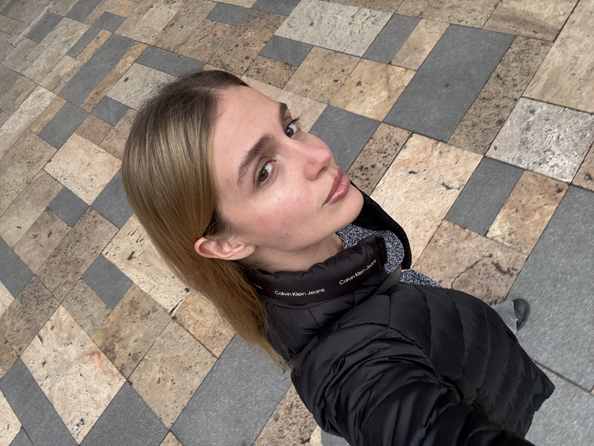A medium-shot angled upward photograph shows a fair-skinned woman with brown eyes and light brown hair wearing a black Calvin Klein Jeans puffer jacket. She is standing on a sidewalk paved with patterned rectangular stones of grey and beige colors. The photo appears to be taken in Tbilisi, Georgia, judging by the distinctive paving style typical of the city.