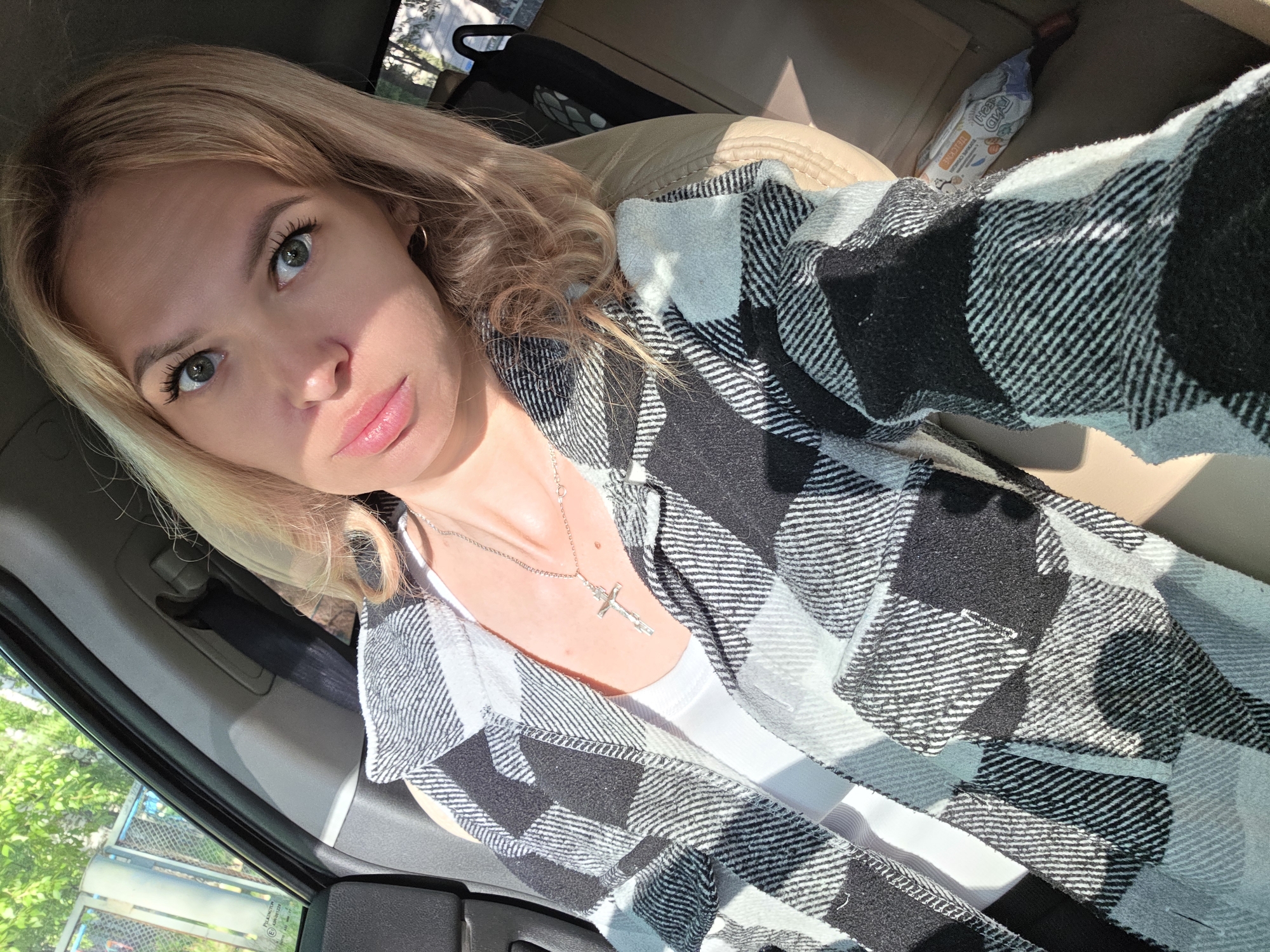 A fair-skinned woman with blond hair and green eyes is visible in a close-up photograph. She wears a silver necklace with a cross pendant and a white tank top under a black and white checkered flannel shirt. The setting appears to be inside a car, based on the presence of a door frame and window. The overall lighting indicates a bright, possibly sunny day. There is a child's snack in the back. There is no specific information in the image that indicates that the location is Chaikovsky, Russia.