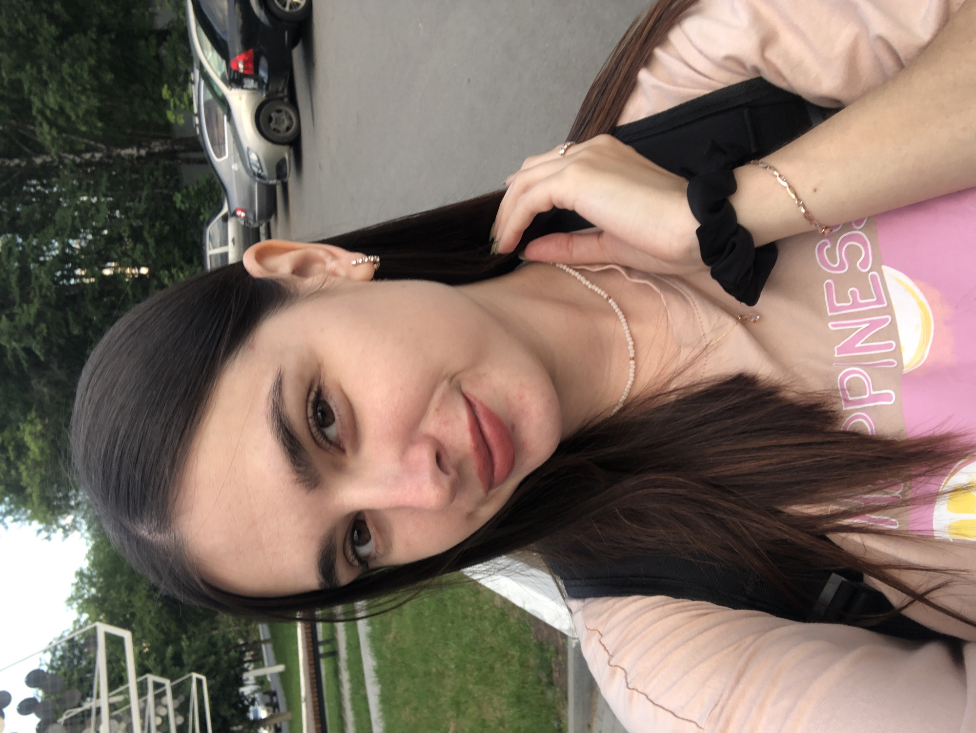 A young woman with long, dark brown hair and fair skin is taking a selfie outdoors. She is wearing a light pink t-shirt with a graphic print and a black backpack. She has on a delicate pearl necklace, small hoop earrings, and a ring on her right hand. Her left hand is touching her hair near her neck, and she is wearing a black scrunchie on her left wrist and a bracelet on her right wrist. The background shows parked cars and trees, suggesting an urban or suburban setting. Based on the style of the cars and the overall environment, this scene is likely located in Chaikovsky, Russia.