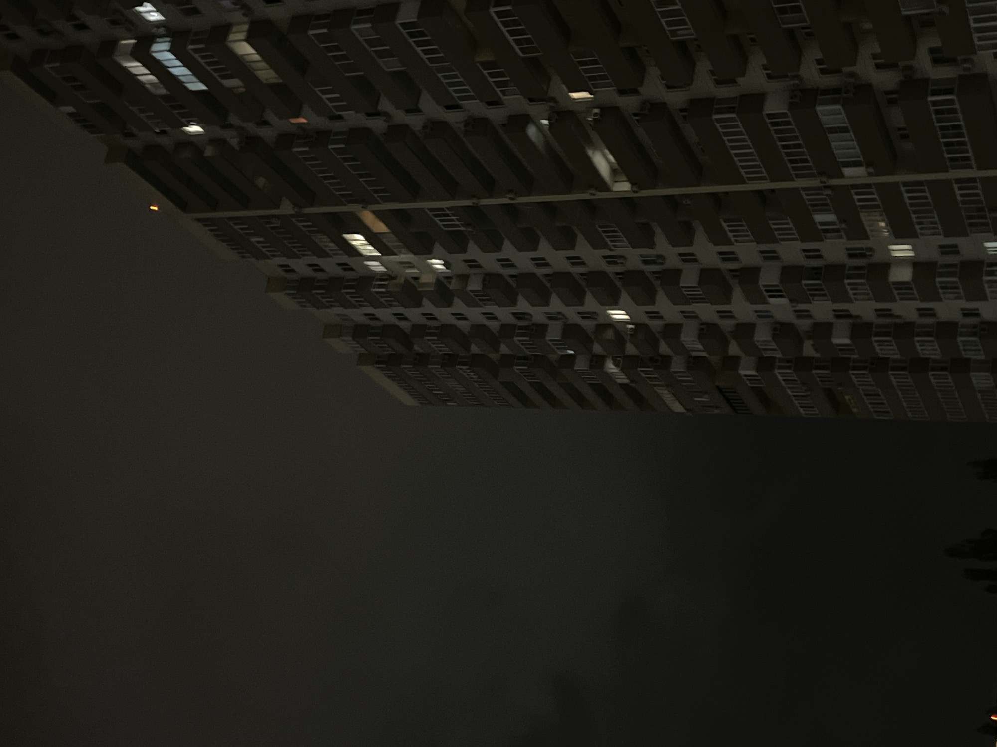 A tall, modern high-rise building dominates the right side of the frame, standing against a dark, overcast night sky. The structure features numerous stacked rectangular sections, likely balconies or window bays, arranged in a repetitive, modular pattern. Many of the windows are illuminated with warm, yellow, and cool white lights, indicating activity within the building. The left and upper portions of the image are filled with the deep black of the night sky, which appears hazy or misty, devoid of stars.
A small, distinct orange light, possibly a distant aircraft or another light source, is visible in the middle-left area of the dark sky, appearing separate from the building. At the very bottom right corner, indistinct dark shapes suggest the presence of foliage, possibly trees. The scene depicts an urban environment at night, emphasizing the scale of the building and the quiet atmosphere. The location context provided is Krasnodar, Russia. No visible text is present in the image.