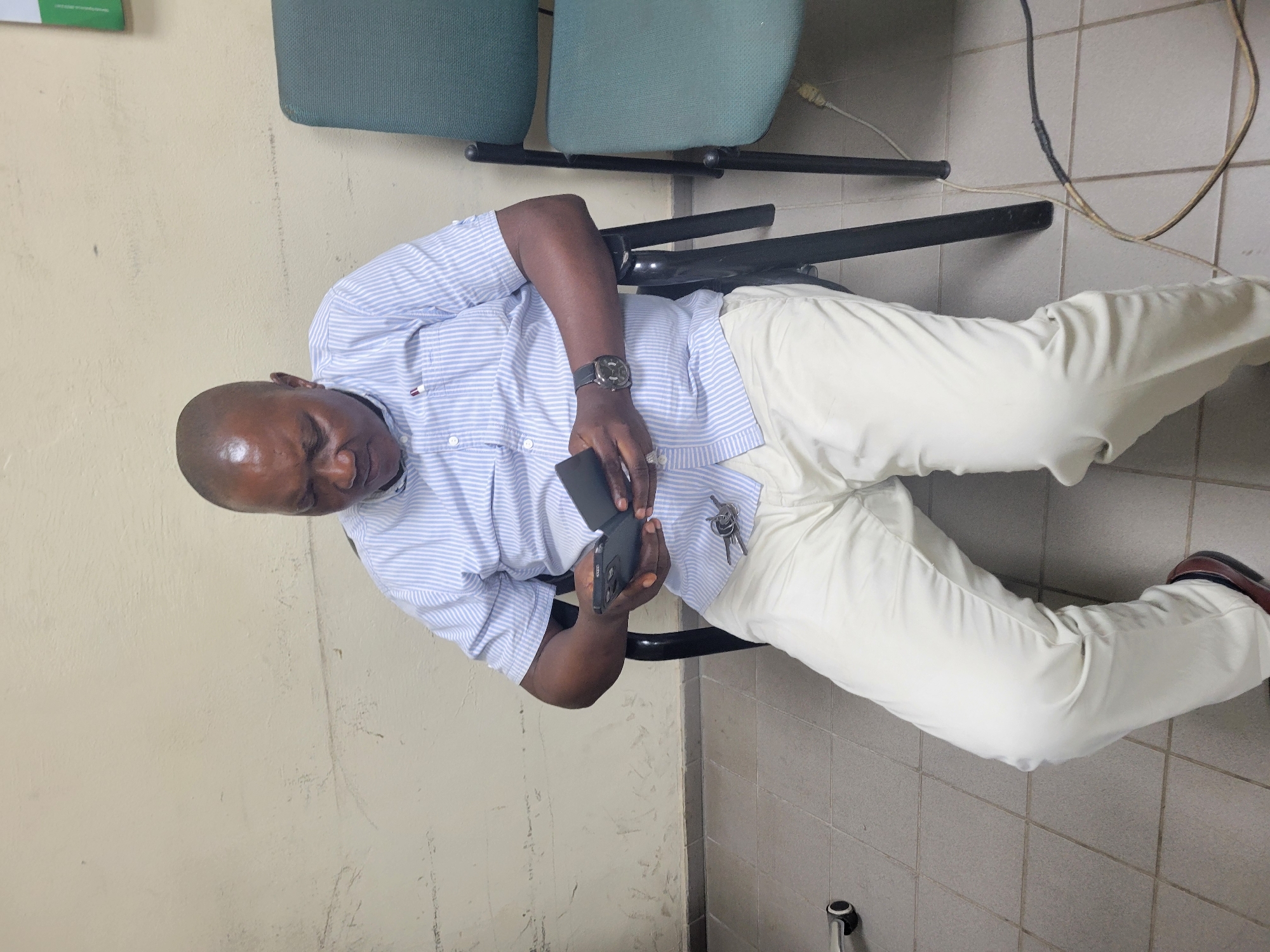 A man is depicted indoors, seated on a chair and engrossed in his smartphone, which he holds in both hands. He appears to be of African descent, with a bald head, and is dressed in a light blue and white striped long-sleeved button-down shirt, cream-colored trousers, and dark shoes. A black watch with a round face adorns his left wrist, and a set of keys hangs from his right hip area. A small, indistinct emblem, possibly a brand logo with red, white, and blue colors, is visible on the left chest area of his shirt.
The setting suggests an indoor institutional or commercial environment, potentially an office or waiting room in Abuja, Nigeria. Behind the man are two chairs with green upholstered seats and black metal frames. The background on the left features a plain, light-colored wall, while the floor on the right is covered with light grey tiles. Several cables and wires are visible on the tiled floor. The lighting is even and ambient, typical of an indoor space, and no specific time of day is indicated. The man's expression is neutral and focused on his device. No other people or significant textual elements are visible in the scene.