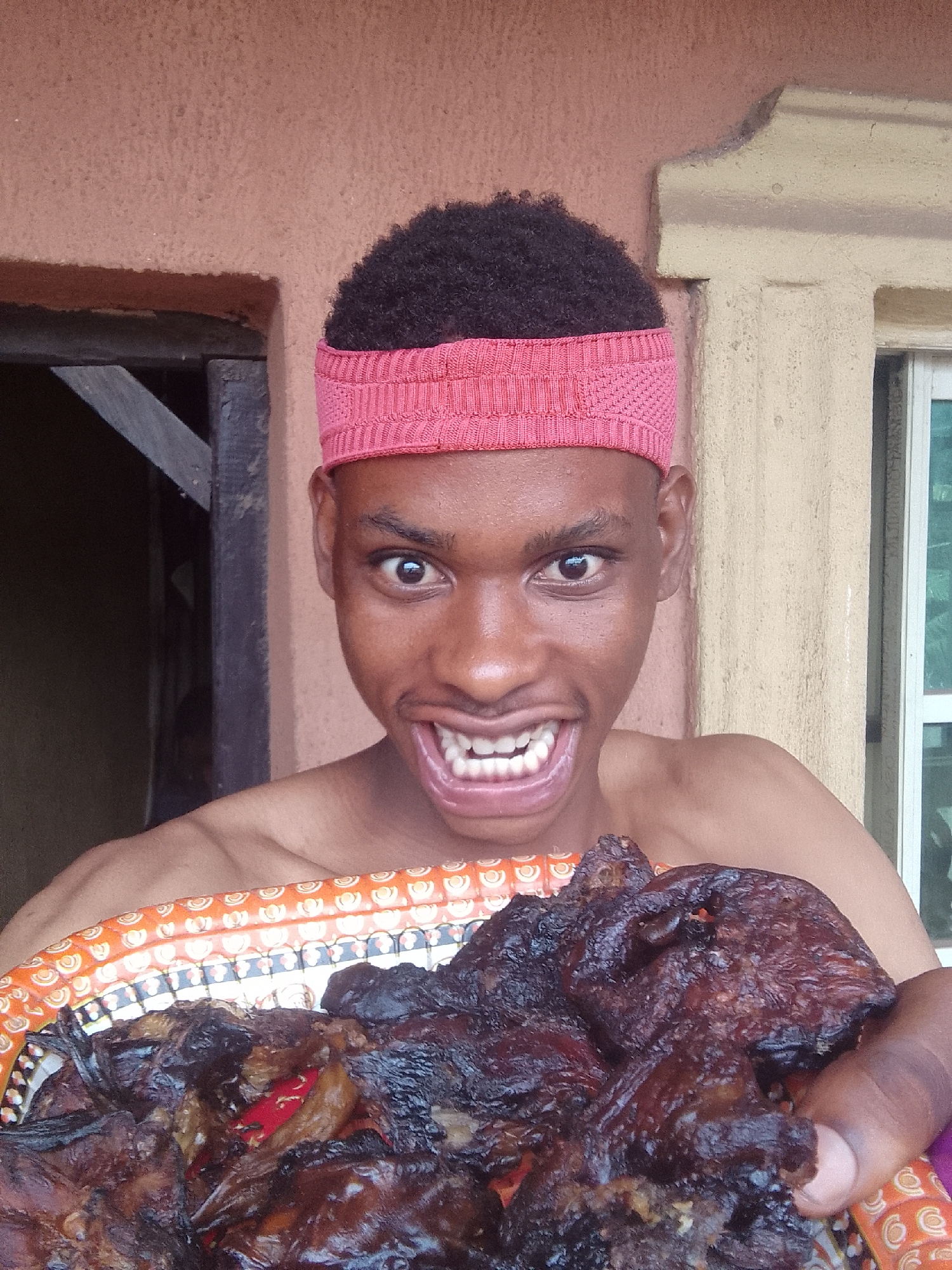 A young man with dark skin, short curly hair, and a bright pink knitted headband is depicted from the chest up, holding a large platter of food. He is shirtless and directly faces the viewer with an exaggerated, wide-eyed, toothy grin that conveys excitement or humor.
The platter he holds is filled with dark, heavily cooked, and somewhat charred pieces of meat, suggesting it has been deeply roasted, smoked, or fried. Some reddish-orange pieces, possibly peppers or other ingredients, are visible amidst the meat. The platter itself has a decorative orange rim with a repeating pattern of small white and black shapes.
The background consists of architectural elements. To the left of the man, a dark doorway or opening with visible wooden beams is present against a textured, light reddish-brown wall. To his right, a different section of wall, colored yellowish-beige with decorative molding, is visible, along with a window featuring light-colored frames and what appears to be green-tinted glass. The scene is brightly lit, indicating daylight. No visible text is present in the image.