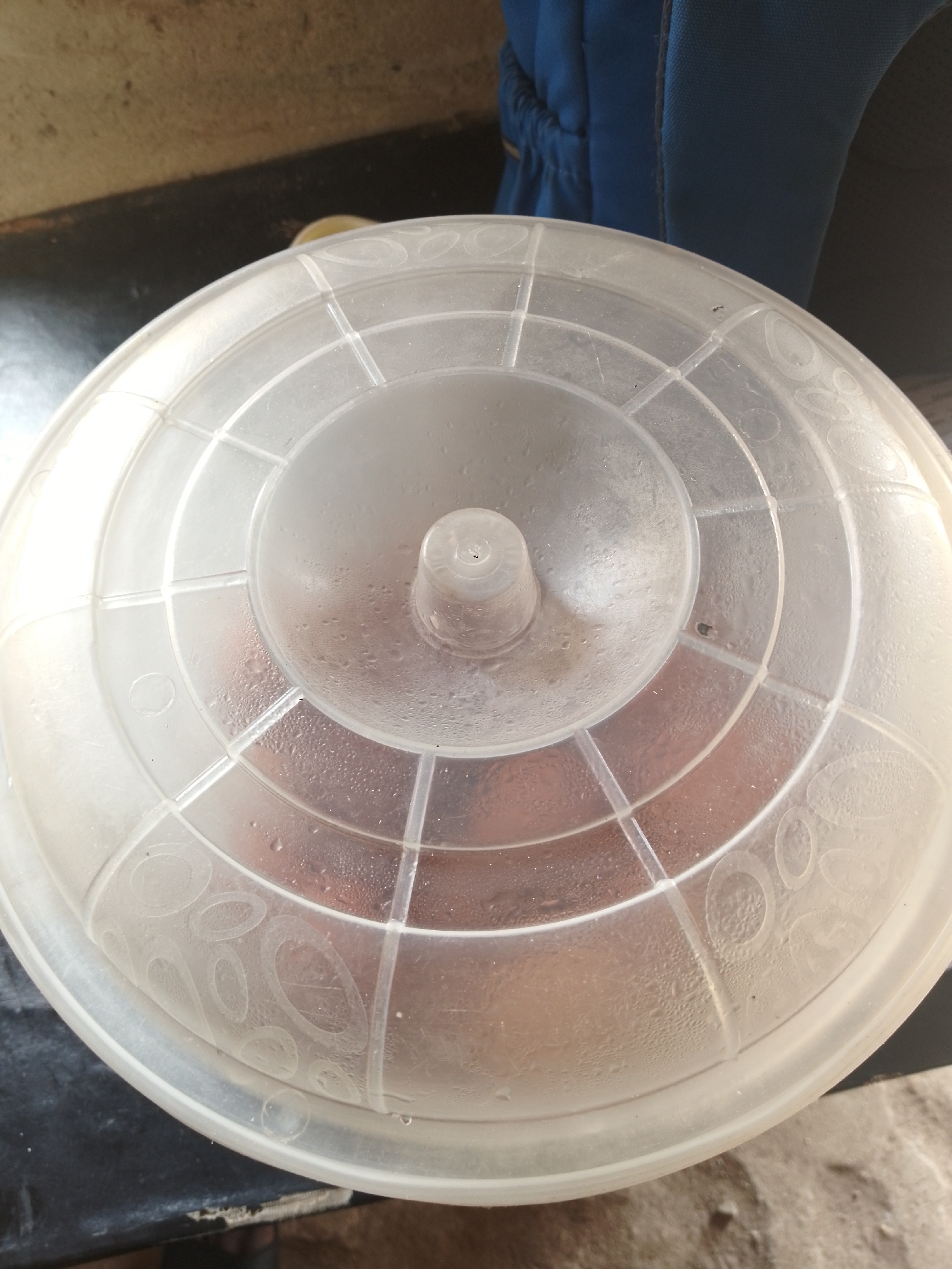 The image shows a close-up, top-down view of a translucent plastic lid covering a food container. The lid has a concentric ribbed design and a raised central knob. Condensation droplets are visible on the interior surface of the lid, obscuring a reddish-brown substance within the container. The lid appears to be made of a flexible plastic material, possibly polyethylene. In the background, a portion of a dark blue fabric item, potentially a bag or clothing, is visible. The lid is resting on a dark, textured surface, possibly a countertop or table. The lighting suggests it is daytime. The scene is located indoors, in a simple, possibly domestic setting. There are no people or visible action in the image. The image is taken from a high angle, looking directly down onto the lid.