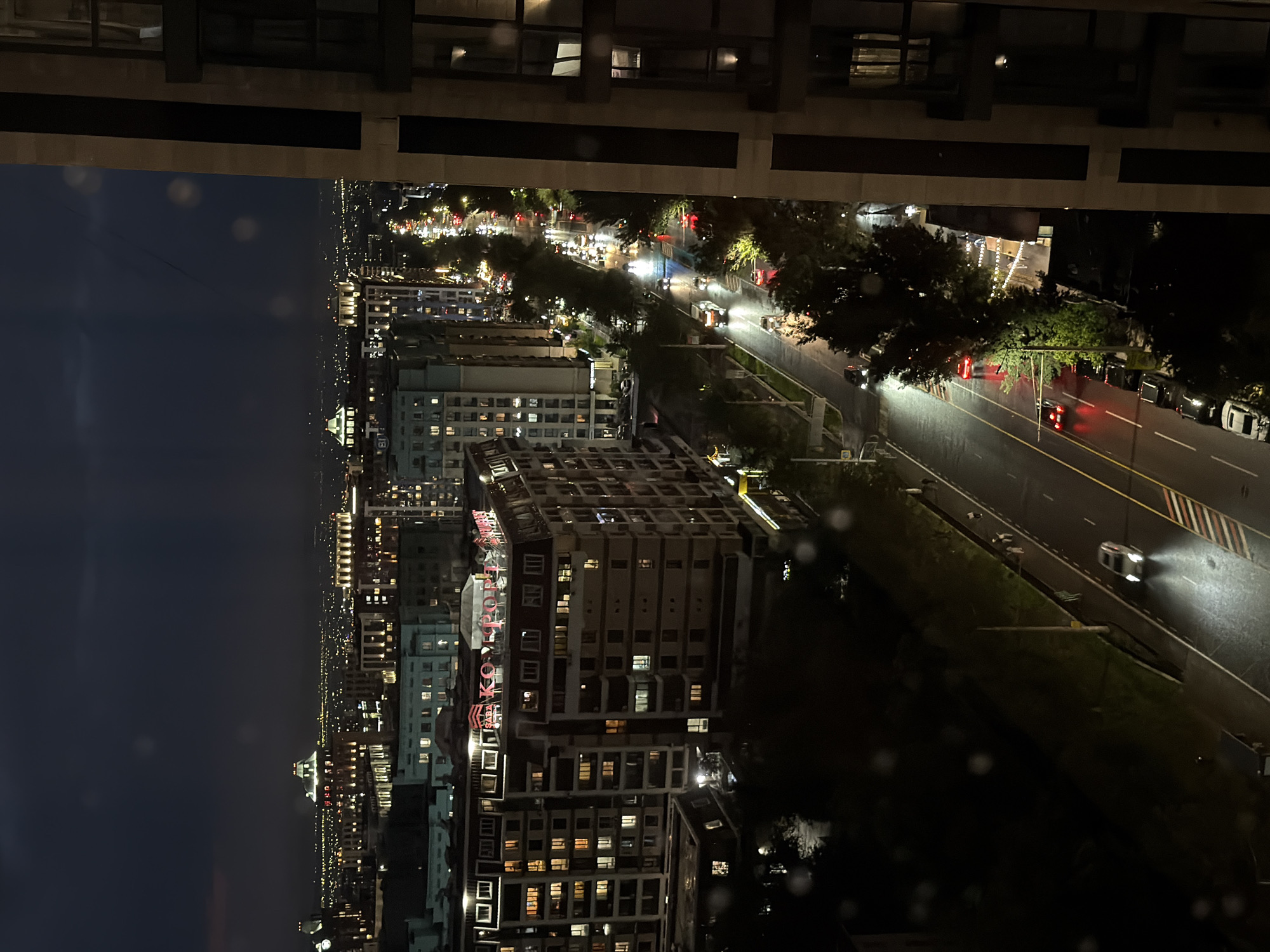 A nighttime high-angle view shows a street scene in Almaty, Kazakhstan. Buildings with illuminated windows are seen to the left of a wide street with traffic lights. Cars and trucks with illuminated headlights and taillights move on the right side of the road. Trees line the side of the road between the buildings and the traffic. In the distance, additional buildings are visible under a dark sky. Above the cityscape, the edge of the building the photograph was taken from is also visible. The lettering "Koktobe" can be seen in red on the side of a building.