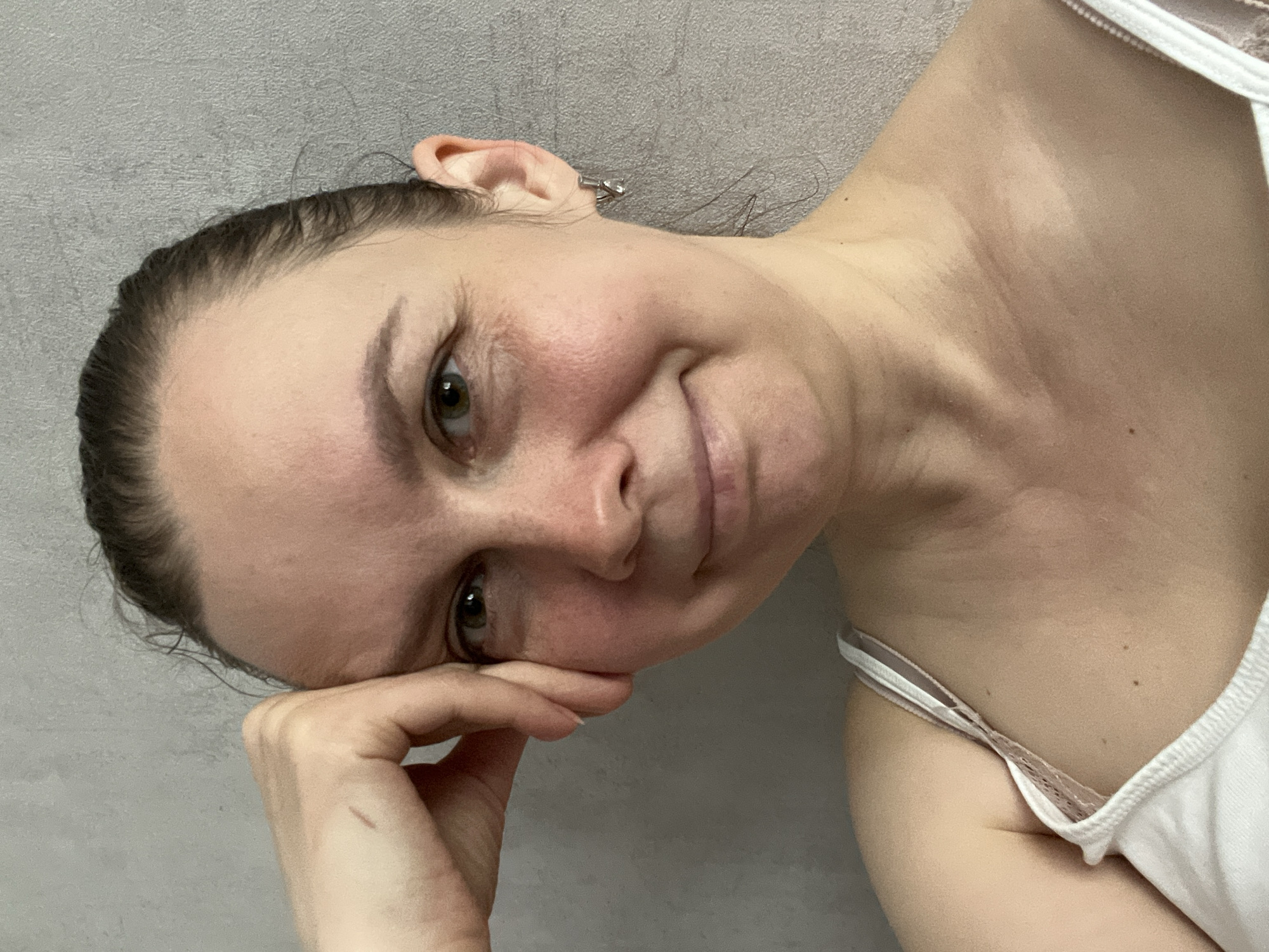 A woman with light skin and brown hair pulled back is looking at the camera with a slight smile. Her head is tilted and resting on her left hand. She is wearing a white tank top. The background is a plain gray wall. The photograph does not contain information to place it in Izhevsk, Russia, nor does the information seem relevant.