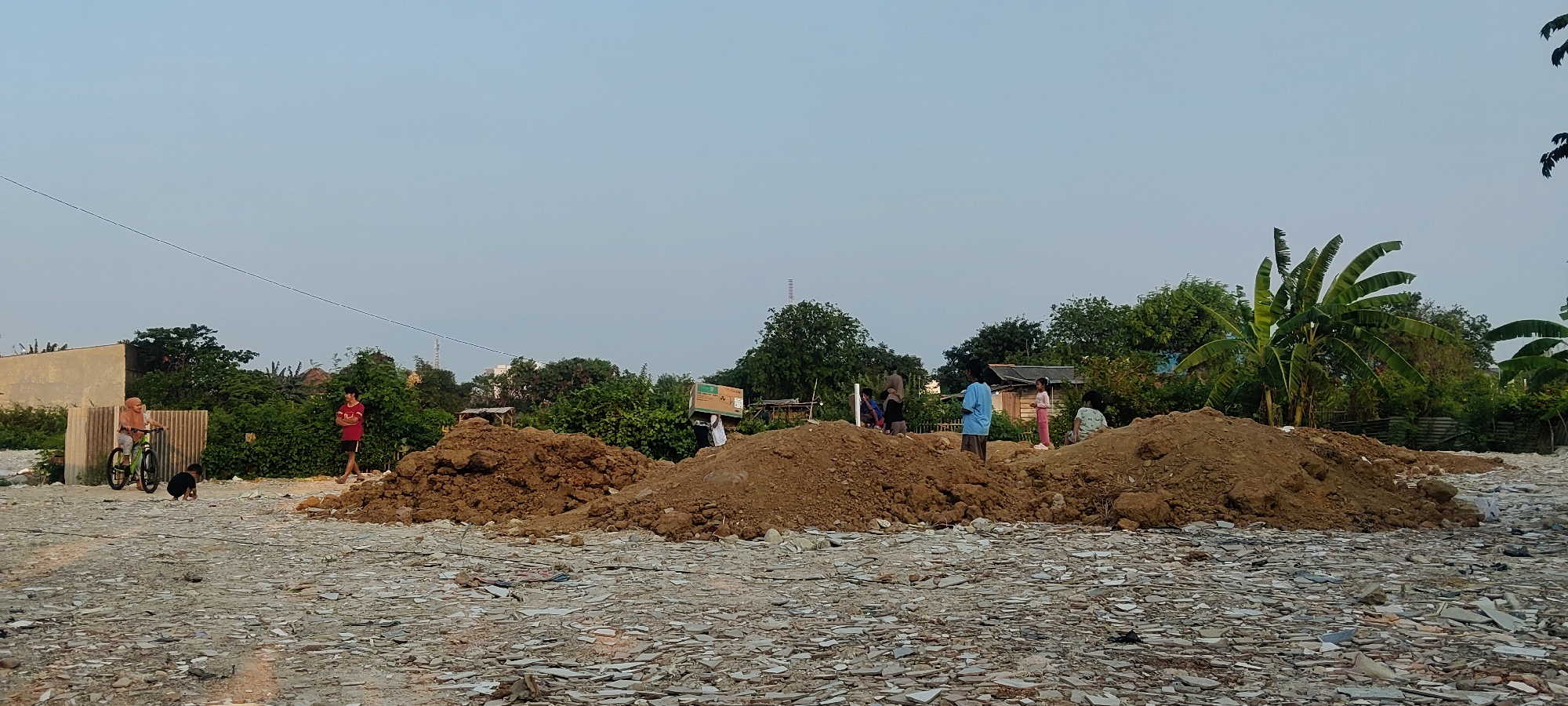 The image depicts an outdoor scene in what appears to be a construction or demolition site in Mekarmukti, Indonesia. The ground is covered with debris, primarily fragments of light-colored material that could be broken tiles or concrete. In the mid-ground, there are several large mounds of reddish-brown soil. A few individuals are visible throughout the scene. On the left, a person wearing a headscarf rides a bicycle. To their right, a young person in a red t-shirt stands. Further to the right, near the soil mounds, a group of people, including children, are present. One person is carrying a large cardboard box on their shoulder. Behind the mounds and people, there is a dense area of green trees and vegetation, with some rudimentary structures, possibly temporary shelters, interspersed. A tall antenna-like structure is visible in the far background, along with some buildings. The sky is a clear, light blue, suggesting daylight. The overall environment appears somewhat unkempt and suggestive of ongoing activity related to construction or land clearing.