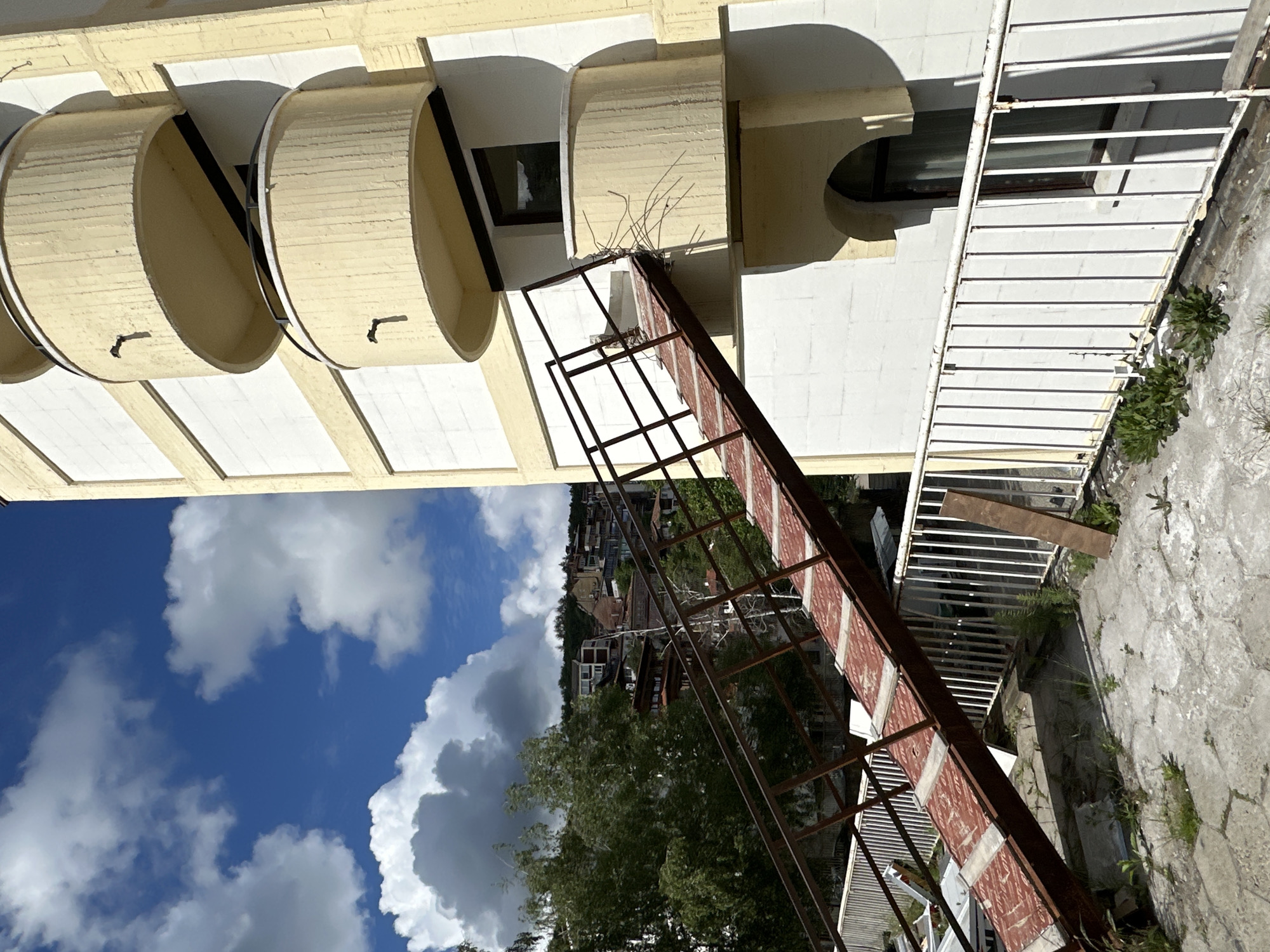 The image depicts an exterior scene in Veliko Tarnovo, Bulgaria, showing an unusual perspective of the built environment. A light yellow building with curved, cylindrical architectural features is situated at the top. Extending from the building is a rusty, metal structure with red-patterned surfaces, resembling a makeshift ramp or decaying staircase. The lower portion of the image features a view of trees, other buildings on a distant hill, and a sky filled with white clouds against a blue background. The image is taken from a tilted angle, creating a disorienting effect where the sky appears to be on the side. A white railing with horizontal bars is also visible in the middle ground.