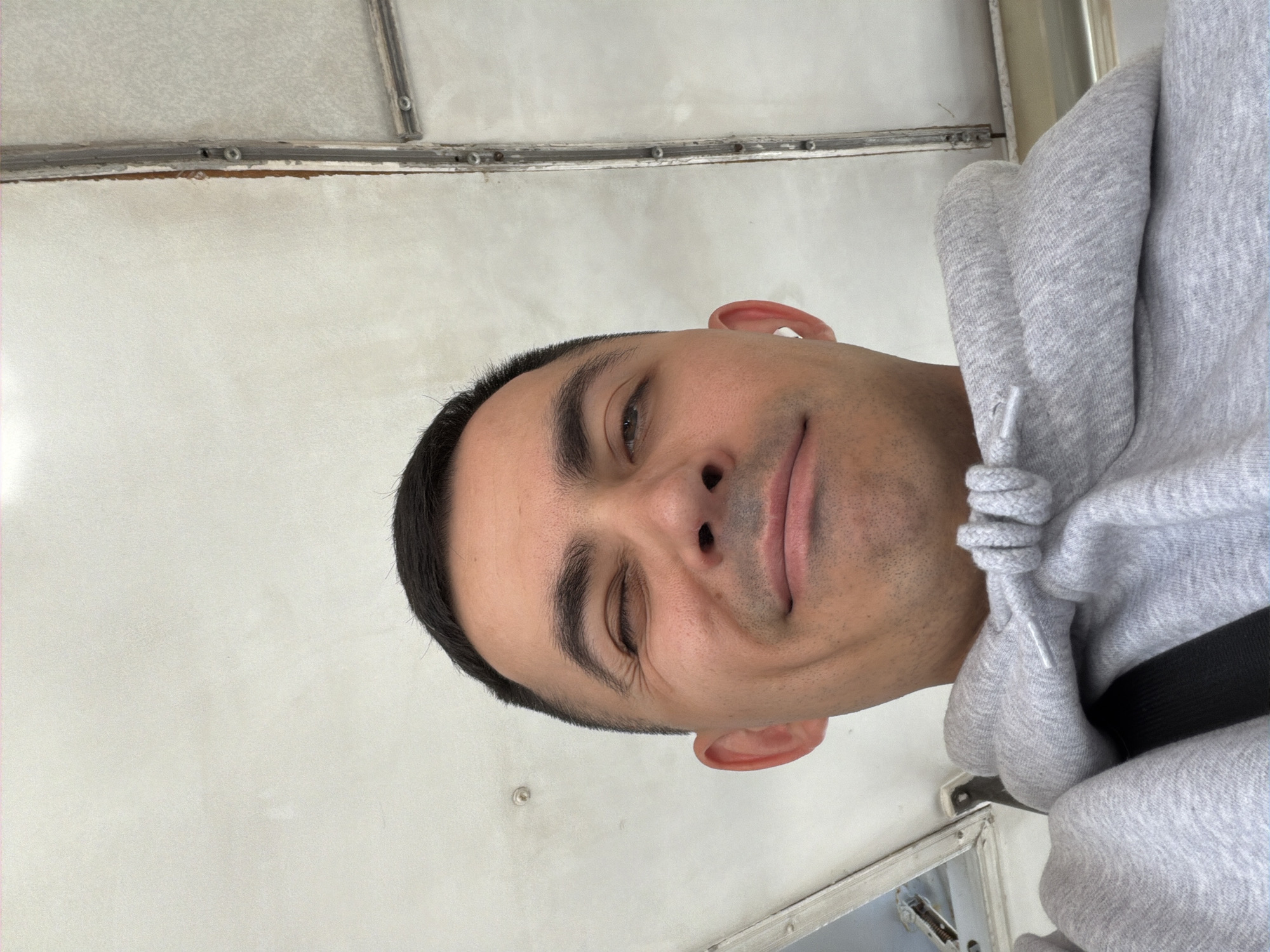 This close-up selfie depicts a man with dark, short hair and a light stubble, wearing a light gray hooded sweatshirt. He has a playful expression, with a slight smirk and his right eye partially closed, suggestive of a wink. A black strap, likely a seatbelt or bag strap, is visible over his right shoulder.
The background consists of light-colored, possibly white or light gray, interior walls that show some wear. Horizontal metallic or wooden strips with visible bolts are present in the upper part of the frame, indicating structural elements. A bright light source or glare is noticeable on the left side of the background. In the lower right corner, a reflective, rectangular panel or opening can be seen. The overall setting appears to be an interior space, possibly within a vehicle such as a bus or train, given the utilitarian nature of the background elements. The image is taken in Izhevsk, Russia. There is no visible text.