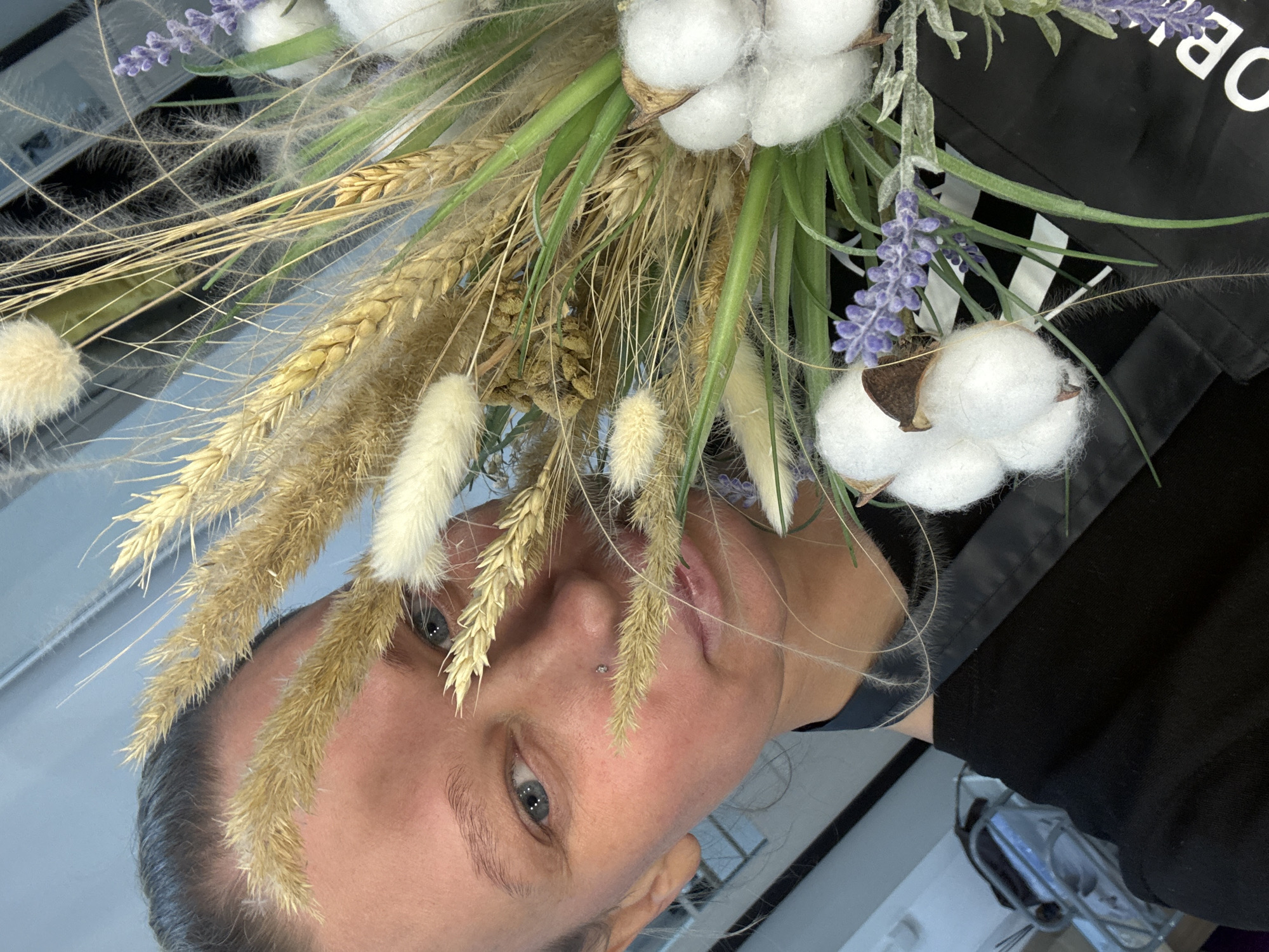 The image, rotated 90 degrees clockwise, captures a close-up of a person's face partially obscured by a dried floral bouquet, set indoors in Perm, Russia. The subject, likely female, has fair skin, light blue/grey eyes, and light brown hair, looking directly at the camera with a neutral expression. A small, sparkling nose piercing is visible.
The prominent bouquet showcases a variety of natural elements: fluffy white cotton bolls, delicate purple lavender spikes, golden wheat stalks, and several types of light beige/white dried grasses with soft, feathery textures, possibly including Lagurus ovatus. Some green foliage is also visible within the arrangement.
The person is wearing a black garment, likely a top or jacket. The background features a reflective surface, possibly a mirror, which shows indistinct reflections of an indoor environment, including a light-colored ceiling or wall and some darker elements. In the upper right corner, on a black background, part of a white uppercase letter "O" is visible, along with a partial "B" or "R," suggesting text like "JOB" or a brand name. The overall impression is a personal portrait featuring a natural, rustic aesthetic.