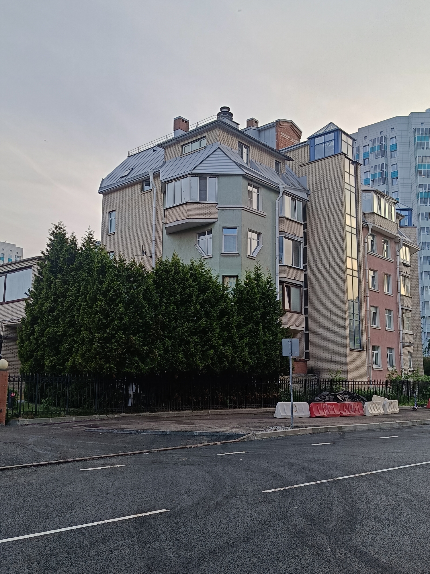 This image depicts a residential street scene on Verbnaia Ulitsa in the Primorsky District of Saint Petersburg, Russia, under an overcast sky.
In the foreground, an asphalt road surface occupies the lower third of the frame, featuring two parallel white dashed lines. A raised curb separates the road from a paved sidewalk on the right. On the sidewalk, a series of five plastic water-filled traffic barriers are arranged horizontally: two white, two red, and one white. Behind these barriers, a pile of dark, irregular debris, possibly asphalt chunks or roofing material, is visible against the fence.
The midground is dominated by a dark green, dense row of columnar evergreen trees, likely Thuja, behind a black metal picket fence. The trees obscure the lower sections of the buildings behind them. To the far left, a glimpse of another building with large windows is visible behind the fence.
The main subject of the image is a multi-story residential building, appearing to be a complex of connected structures, situated behind the trees and fence. This building displays a varied architectural style. The left section is constructed of light-colored brick with a mansard roof featuring a skylight. A section to its right transitions to a mint-green rendered facade with several windows and a bay window. Further right, a section of the building features a prominent vertical glass-encased structure, likely an elevator shaft, constructed with light brick cladding. This section abuts another part of the building with a dusty-pink rendered facade, also featuring multiple windows and a bay window. The building exhibits multiple gray metal downspouts and several brick chimneys on its roofline. The roofs are predominantly gray metal, some with mansard designs.
In the background, beyond the brick and rendered building, a significantly taller, modern residential high-rise is visible. This building is characterized by a light blue and white facade with numerous uniformly arranged windows and balconies.
The sky is a uniform, light gray, indicating overcast conditions, which provides soft, diffuse lighting across the entire scene without distinct shadows. There are no visible people or vehicles in the immediate scene.