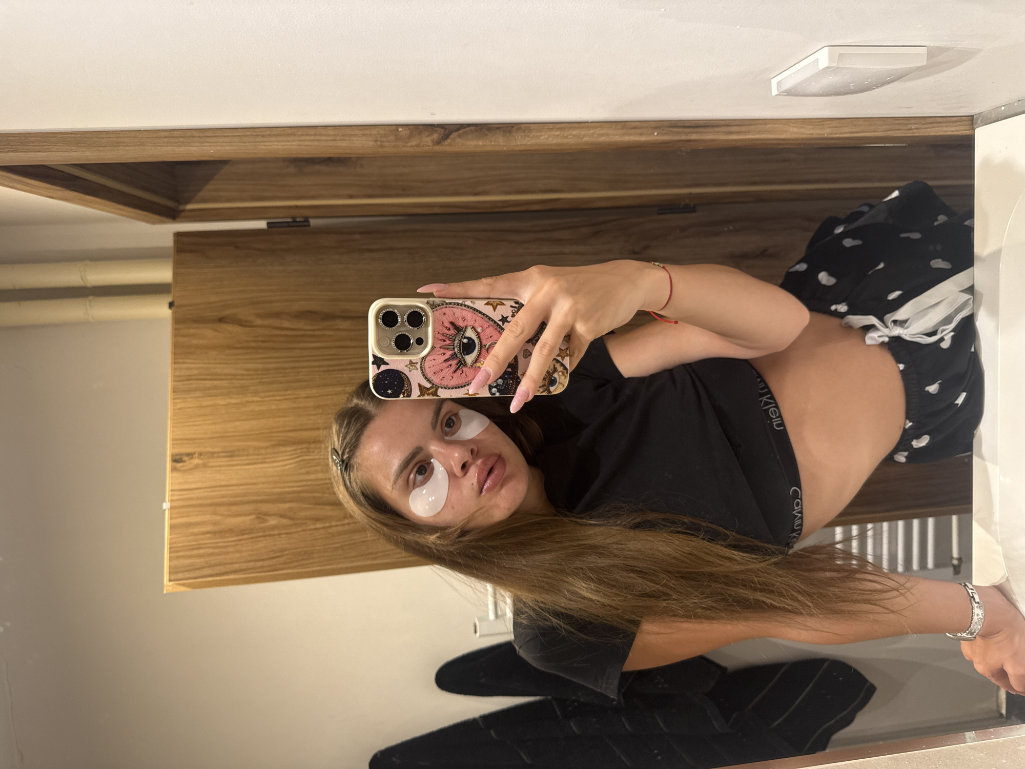 A young woman is captured in a mirror selfie, looking directly into the camera. She has long, light brown hair styled with a hair clip, and distinct under-eye patches are visible below her eyes, suggesting a skincare routine. Her lips appear full and glossy. She is wearing a black crop top, revealing her midriff, and black patterned pajama-style shorts, with the white waistband of "Calvin Klein" underwear or shorts visible.
Her left hand holds a smartphone with an ornate, light-colored case featuring an "evil eye" design and celestial motifs. The phone has a prominent multiple-lens camera system. She wears long, light pink fingernails, a thin red string bracelet on her left wrist, and a silver-toned bracelet with white stones on her right wrist.
The background reflected in the mirror behind her primarily consists of warm-toned wooden cabinetry or paneling. Above this, a light-colored wall and a white, rectangular ceiling fixture are visible. The overall setting appears to be an indoor space, possibly a bathroom or dressing area, characterized by its modern wooden elements and clean design. The image captures a moment of self-care and casual attire.