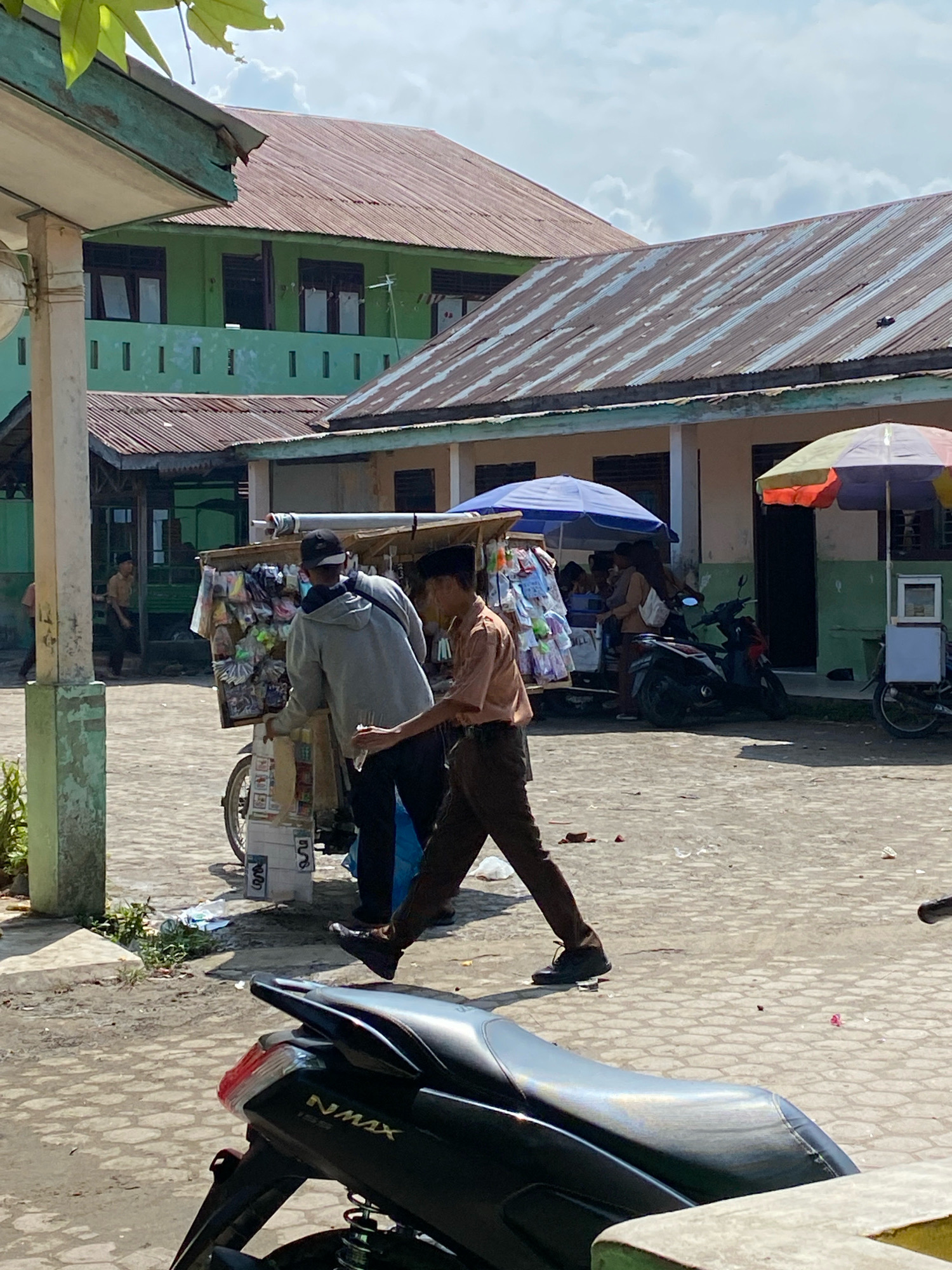 In Karang Gading, Indonesia, on a sunny day, a street scene unfolds with two young men, likely students based on their school uniforms, interacting near a mobile cart selling various goods. One student, wearing a brown shirt and dark pants, is walking away from the cart, holding a clear plastic bag with a white object inside. The other student, in a grey hoodie and black cap, stands behind the cart, which is laden with packaged items. Several motorcycles are parked in the background, and buildings with red metal roofs are visible, suggesting a commercial or school environment. The ground is paved with interlocking concrete blocks, with some scattered debris. The image captures a moment of everyday life in this Indonesian locality.
