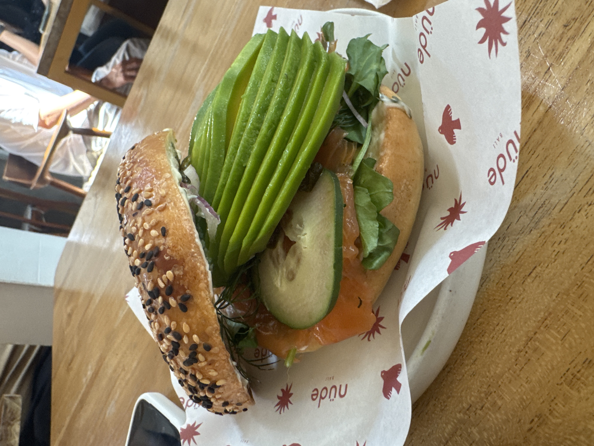 The image depicts a bagel sandwich on a white paper wrapper with a red bird and star pattern, indicating it's from Nüde in Bali, likely in North Kuta, Indonesia, as suggested by the logo on the wrapper. The bagel is topped with sesame seeds, and the filling consists of avocado slices, salmon, cucumber slices, and some greenery, possibly arugula or dill. The sandwich is placed on a wooden table, and a partial view of chairs and other background elements are visible, suggesting an indoor cafe setting.
