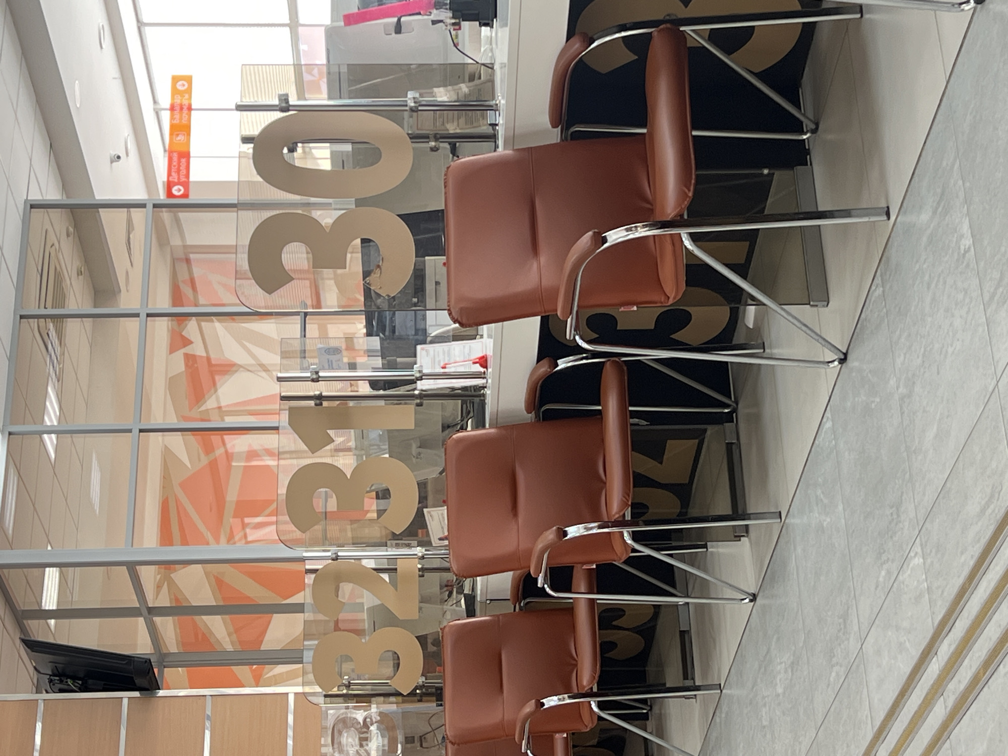 The image shows a waiting area inside a branch of Ak Bars Bank in Kazan, Russia. The waiting area has a glass partition with the bank's phone number "8-800-200-53-03" displayed in large numbers. In front of the partition, there are several brown leather chairs with metal frames arranged in a row. The floor is light gray tile. A sign with text in Russian is visible near the top left.